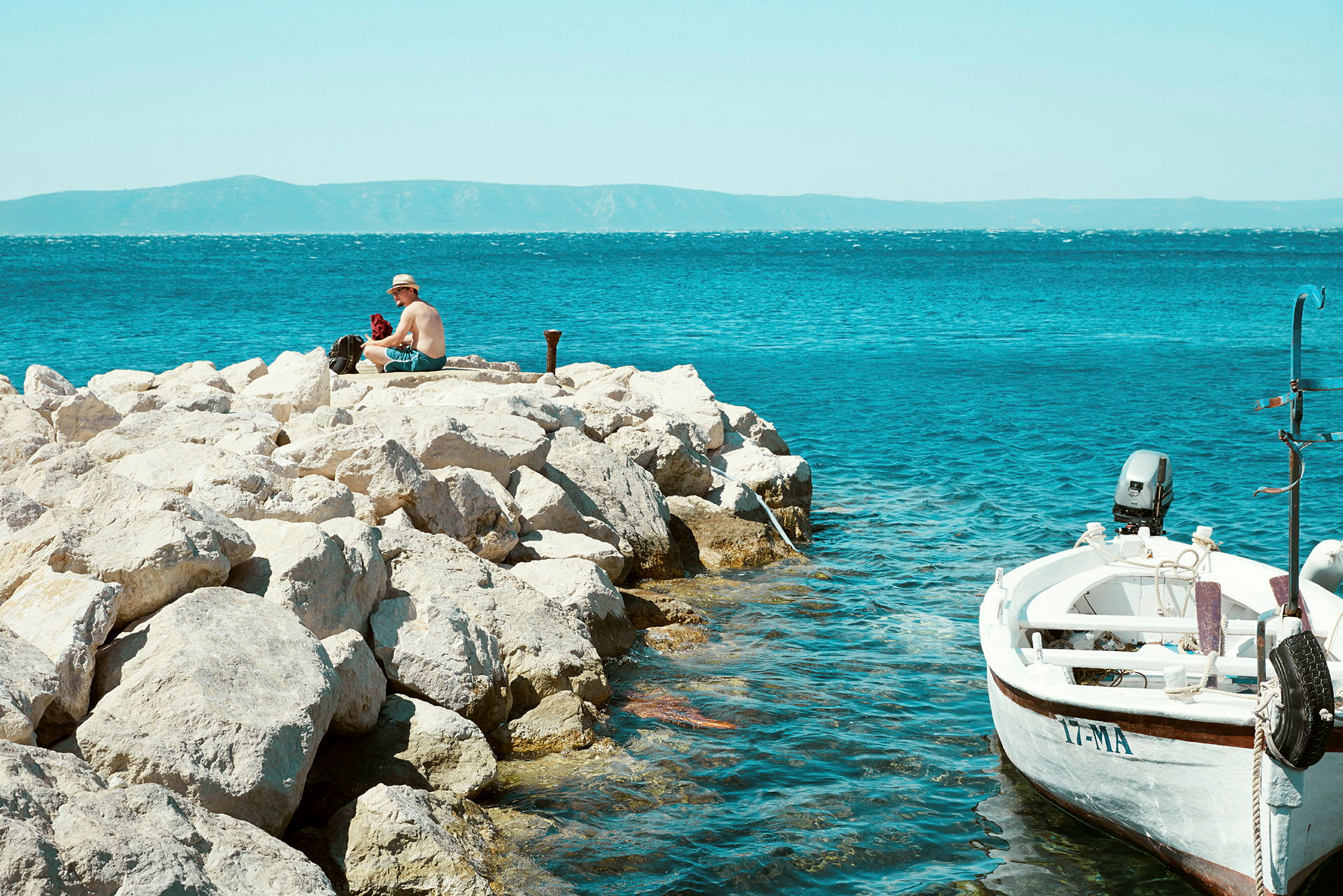 En person sidder på klipper nær havet, med en lille båd fortøjet ved siden af. (AI-genereret tekst)