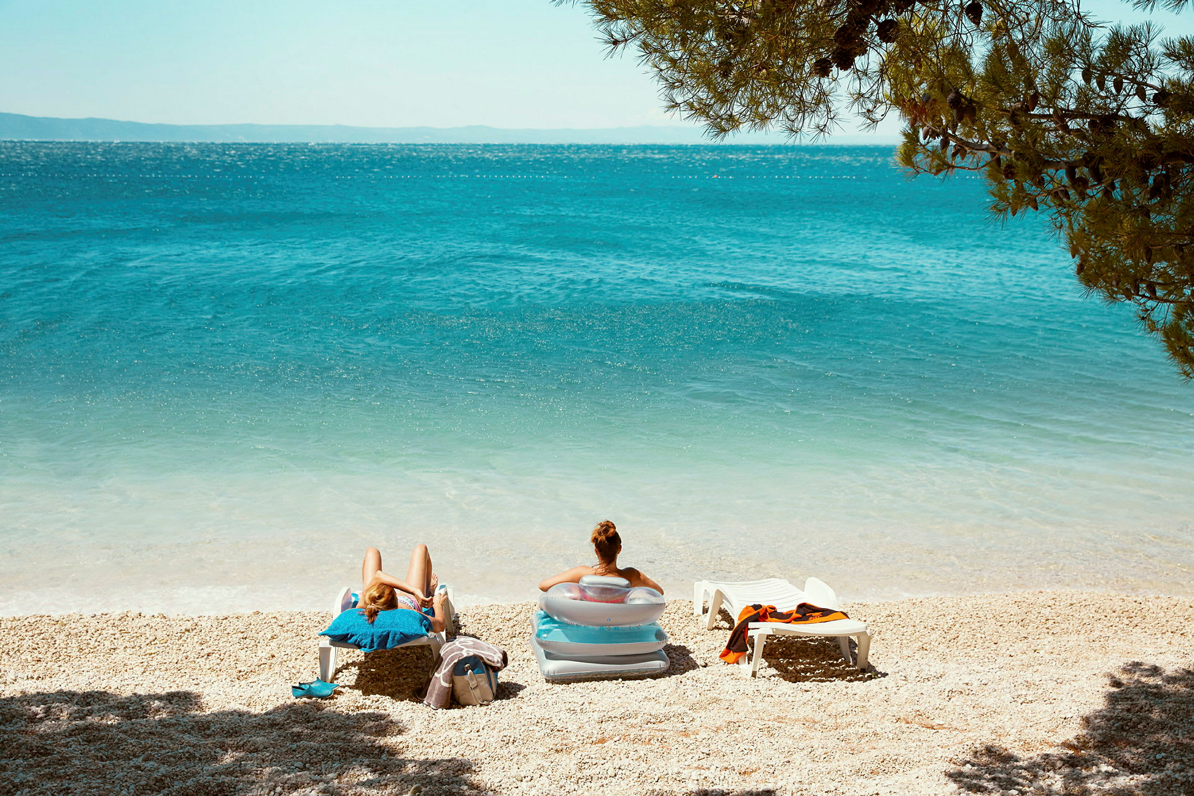 To personer sidder i solsenge på en strand med blåt hav og omgivende grønne områder. (AI-genereret tekst)