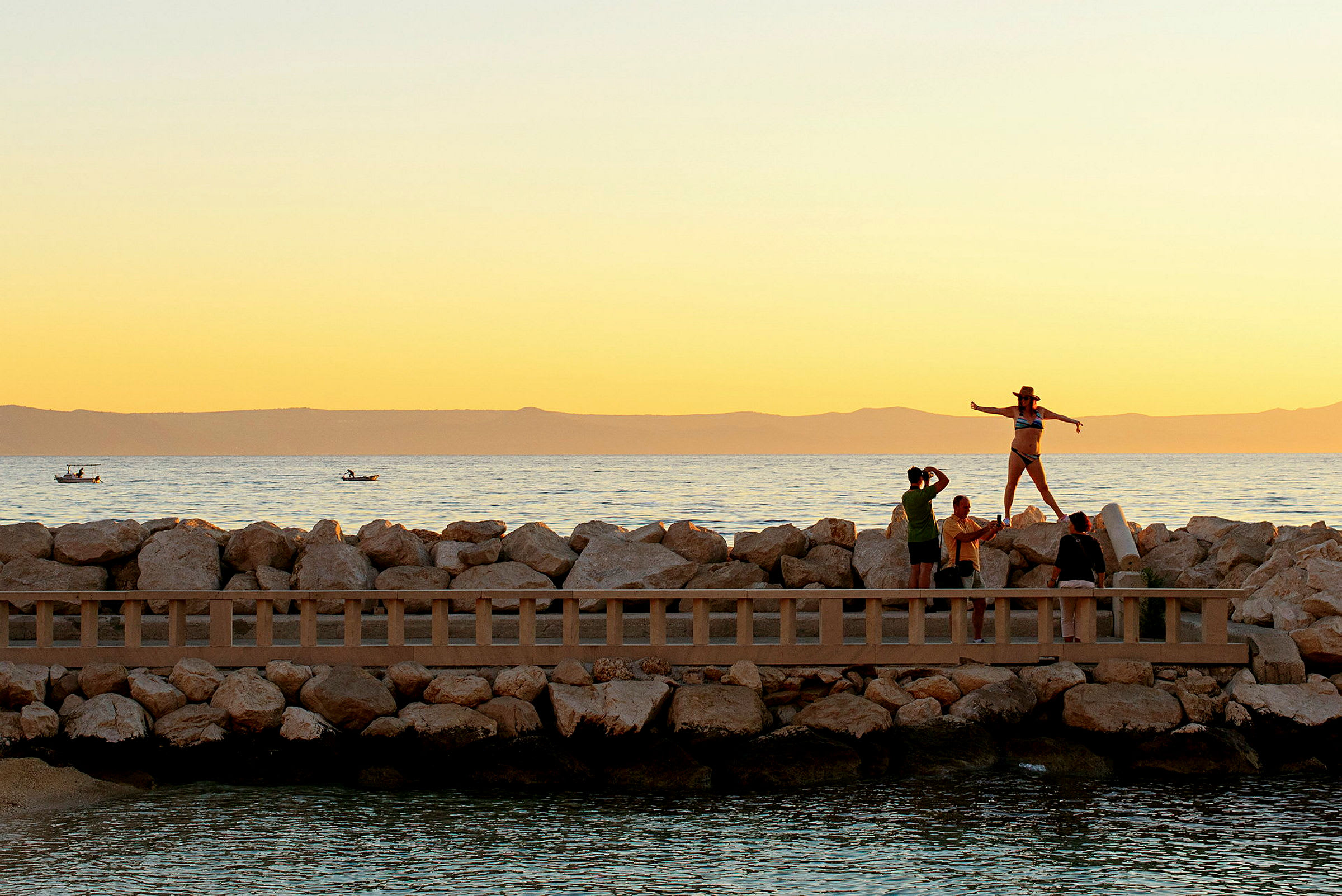 Folk ved en stenmur tæt på havet ved solnedgang. (AI-genereret tekst)