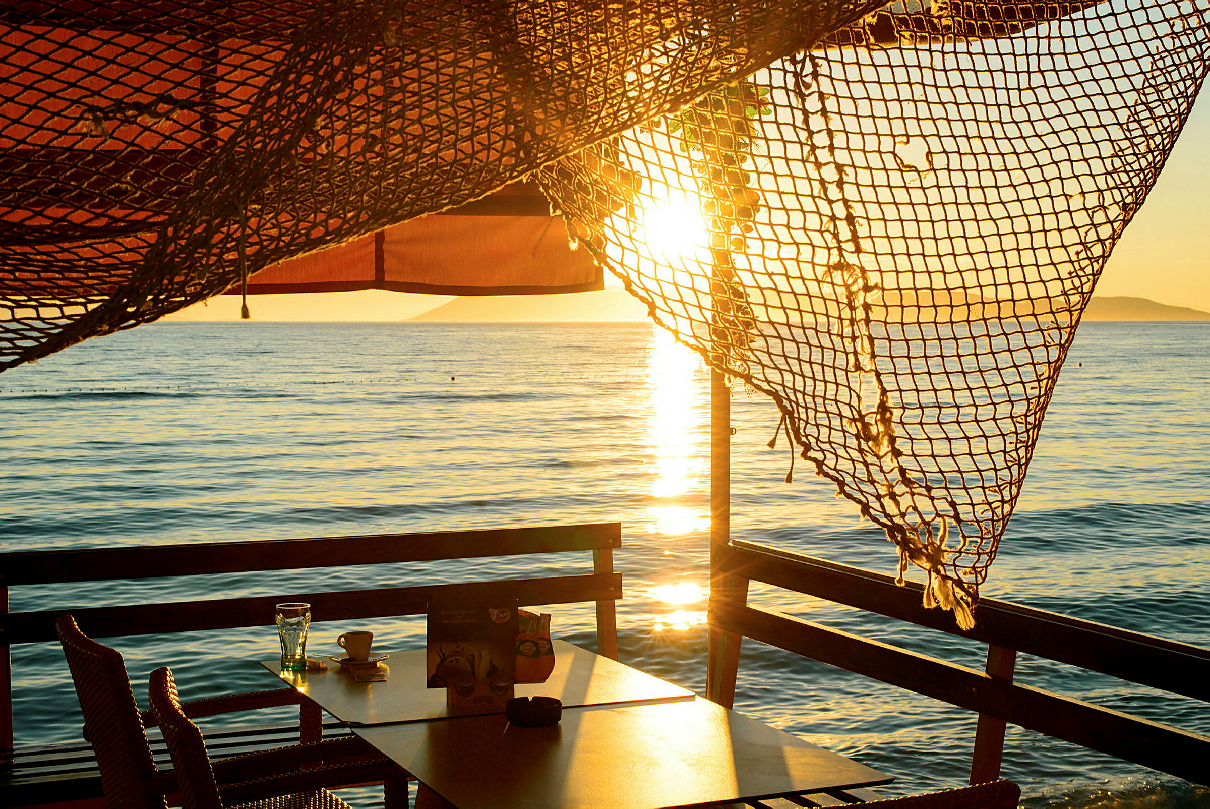 Udsigt over havet fra en udendørs servering med bord og stole, skygge af net og solnedgang. (AI-genereret tekst)