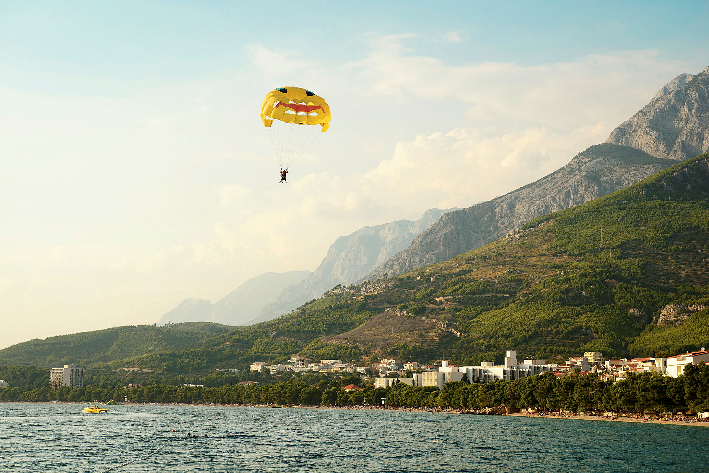En person paraglider med en faldskærm med ansigtsmotiv over en kystlinje med bjerge i baggrunden. (AI-genereret tekst)