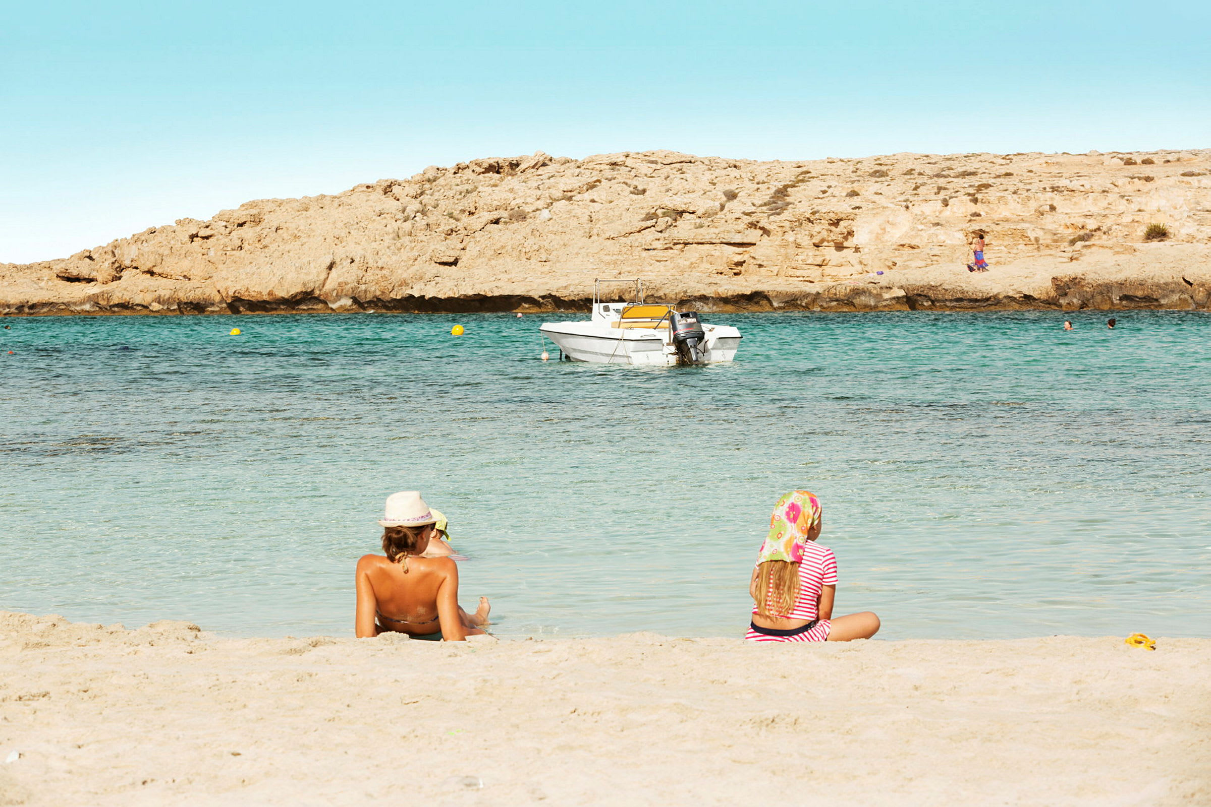 To personer sidder på en sandstrand med havet og en båd i baggrunden. (AI-genereret tekst)
