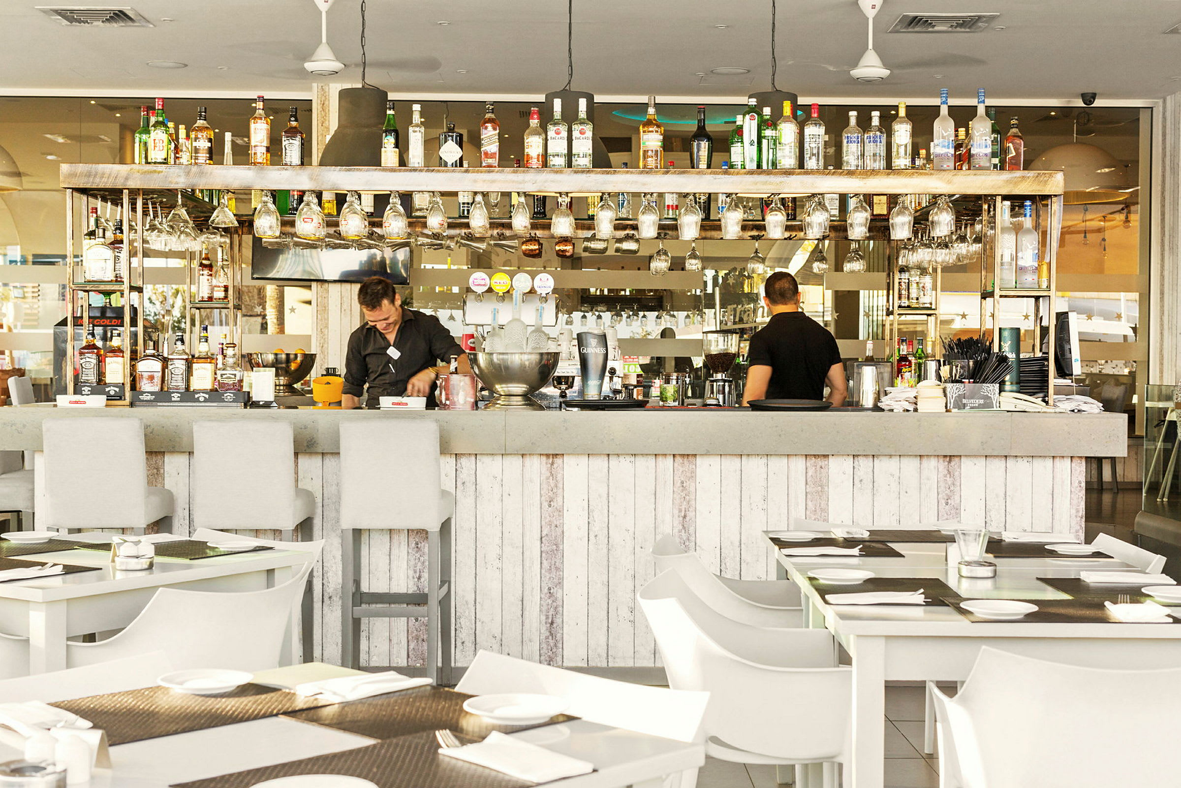 En veludstyret bar med hængende cocktailglas og drikkeflasker på hylden, med siddepladser og dækkede borde foran. (AI-genereret tekst)