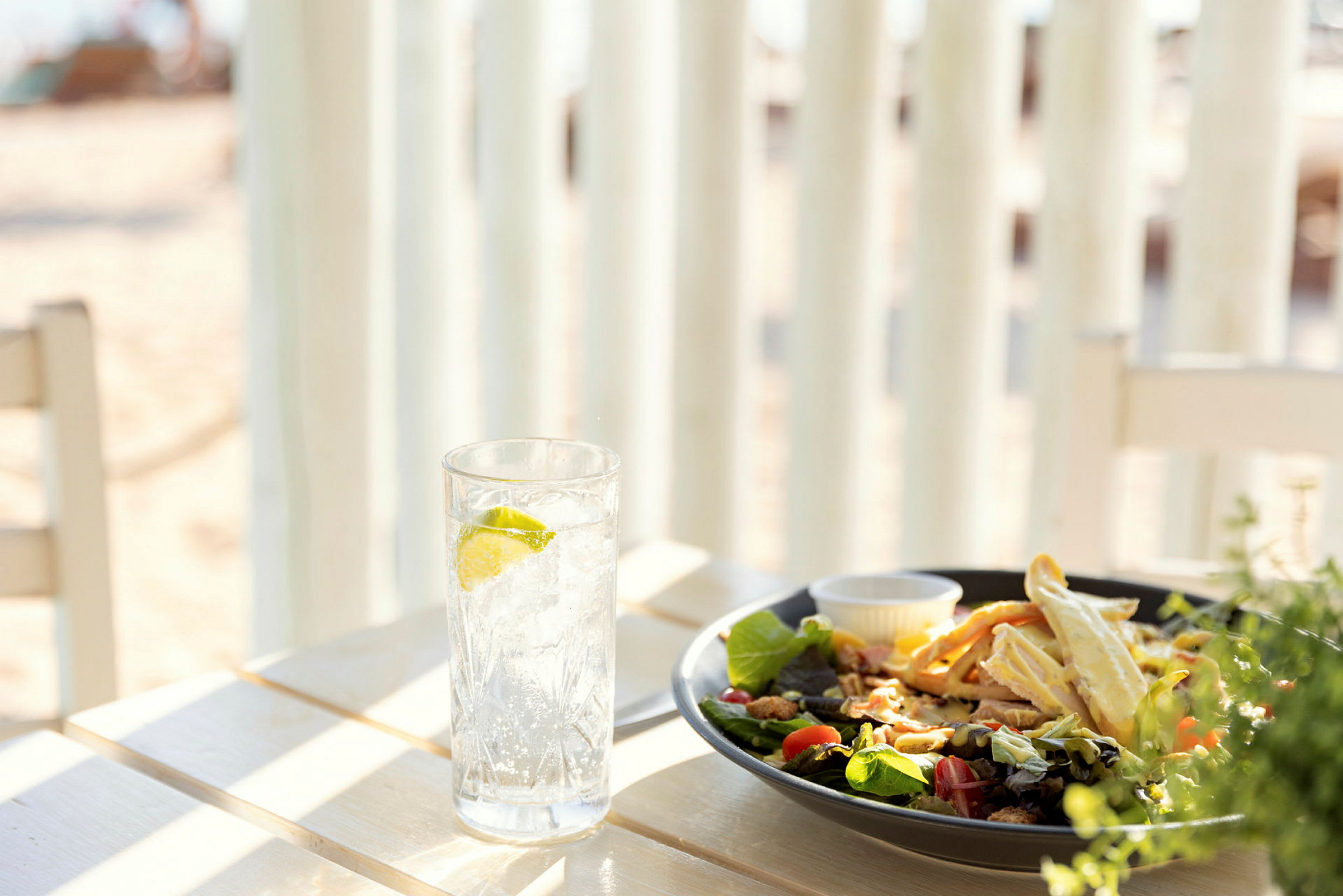 Et bord med et glas vand med lime og en tallerken salat ved stranden. (AI-genereret tekst)