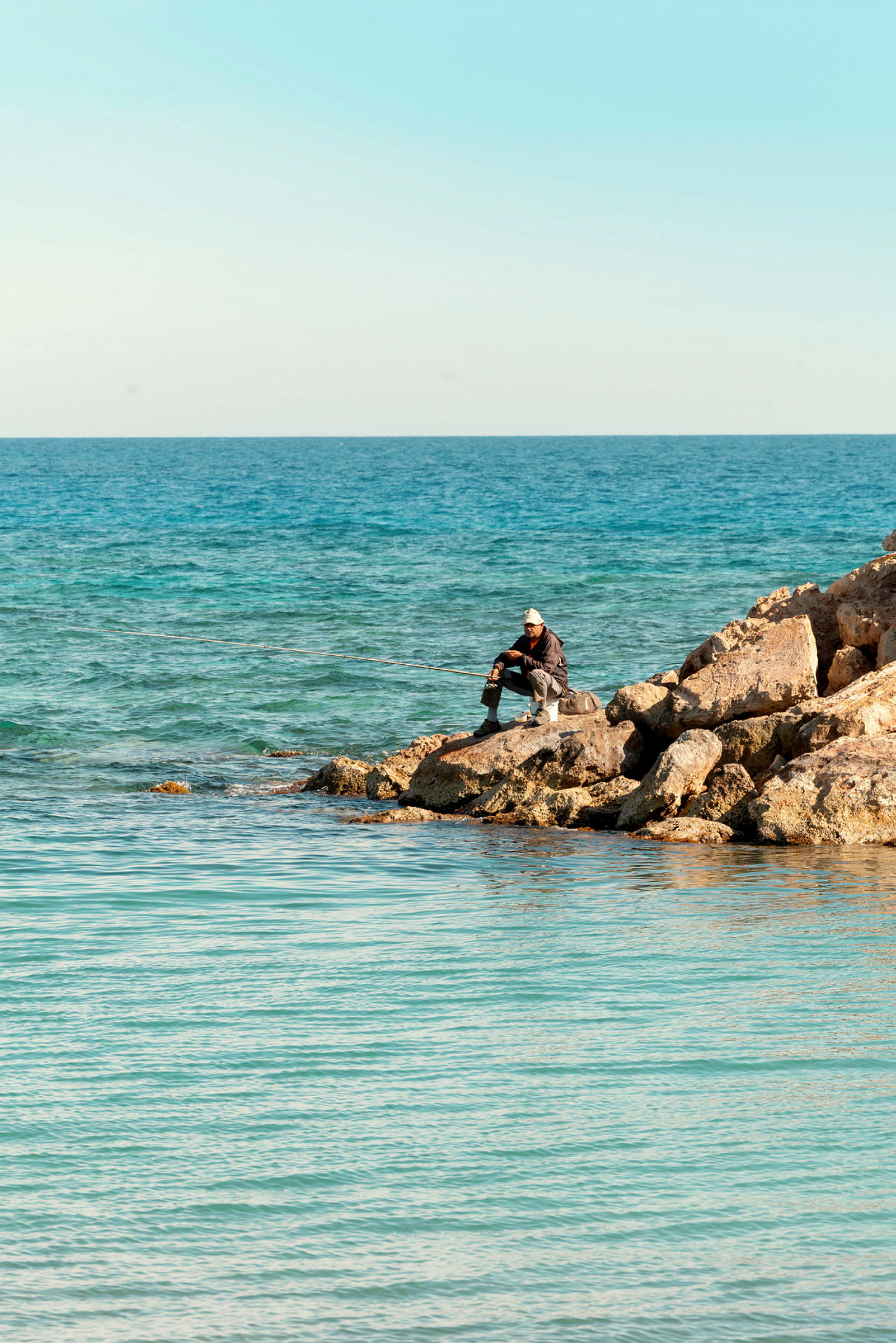En strand med klipper ved havet, med en person der holder en fiskestang. (AI-genereret tekst)