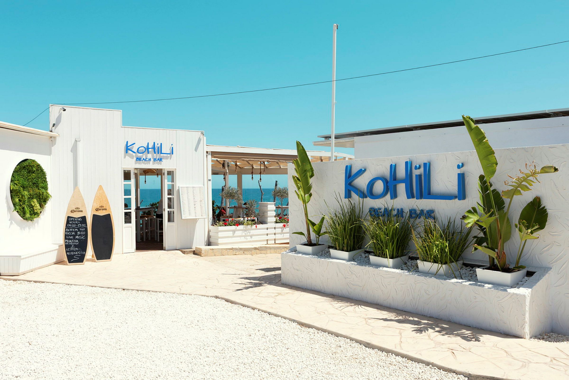 Indgang til Kohili Beach Bar med dekorative planter og en klarblå himmel. (AI-genereret tekst)