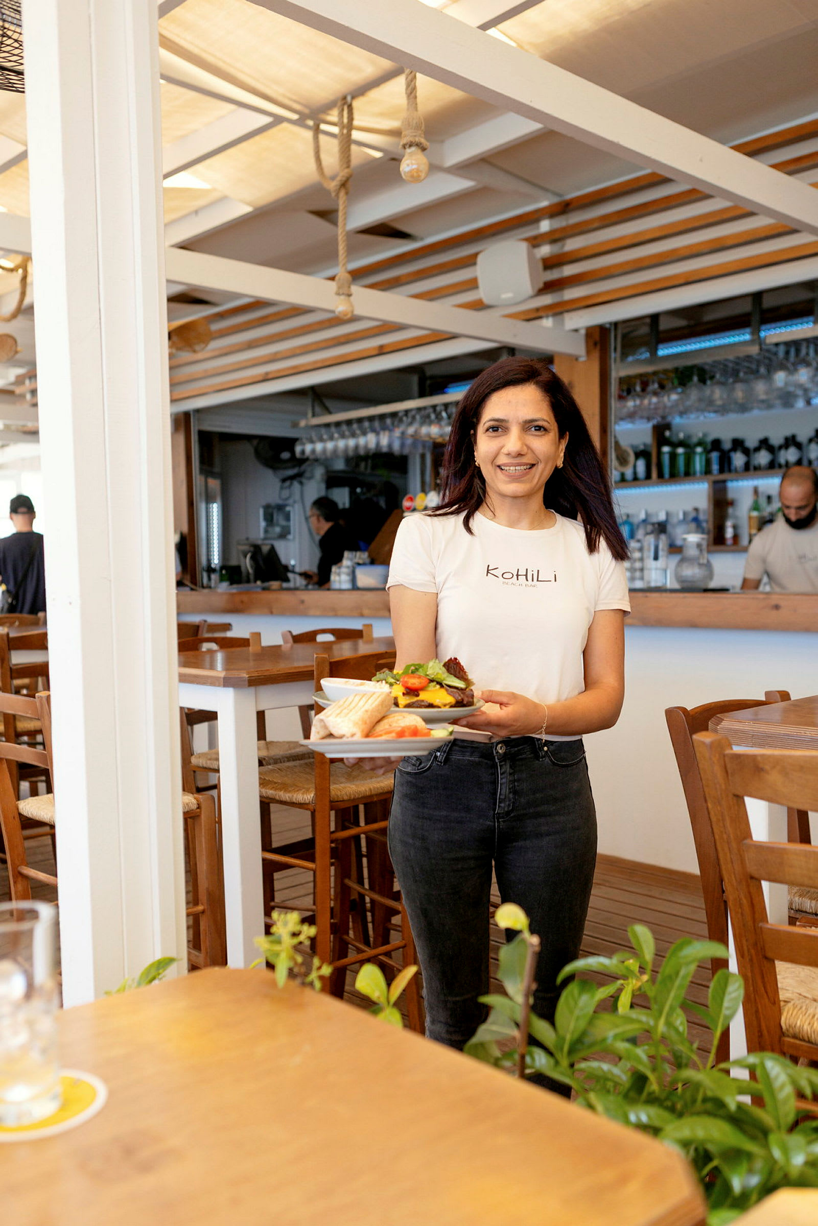 En person holder en tallerken med mad i en restaurant med træinteriør og planter i forgrunden. (AI-genereret tekst)