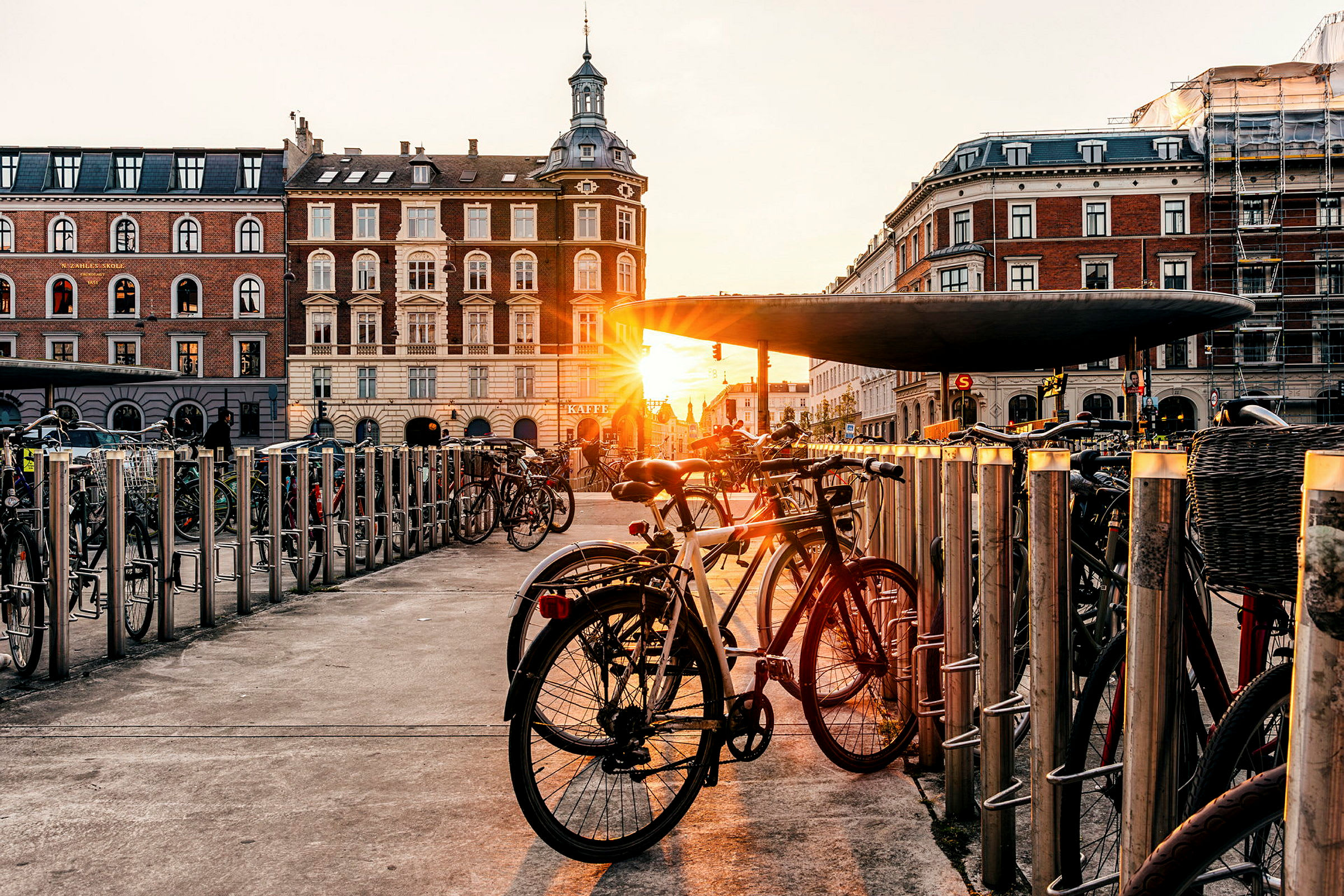 Flere cykler parkeret ved cykelstativer, med bygninger og en solnedgang i baggrunden. (AI-genereret tekst)