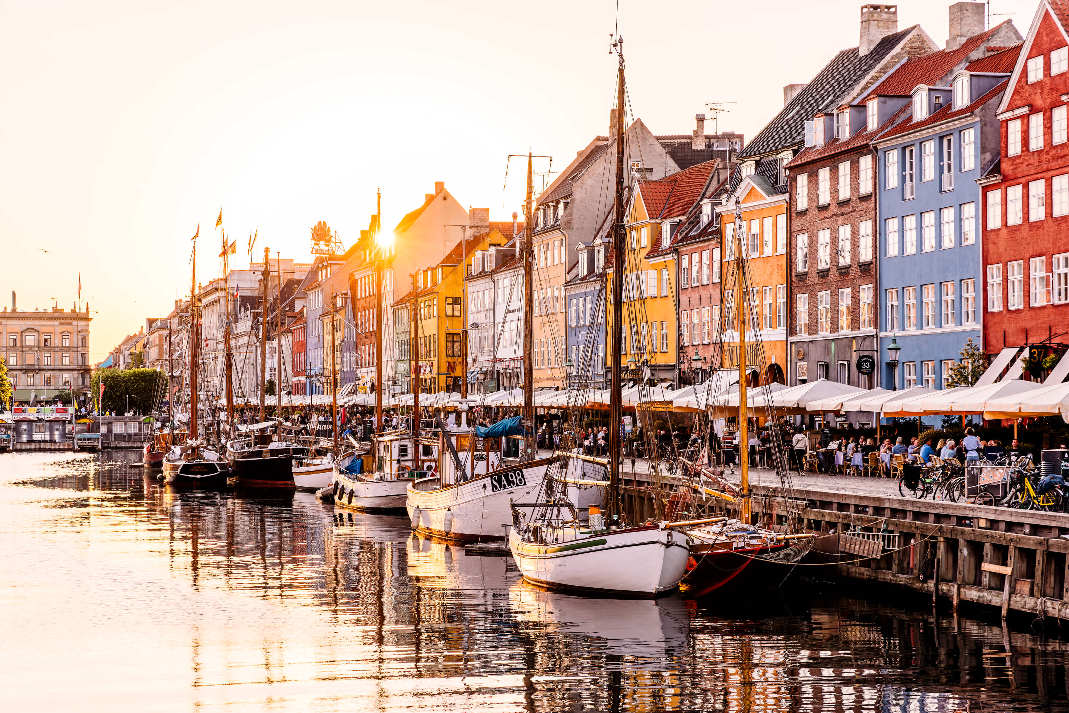 Farverige bygninger langs en kanal med både fortøjet i vandet og mennesker, der sidder ved borde på kajen. (AI-genereret tekst)