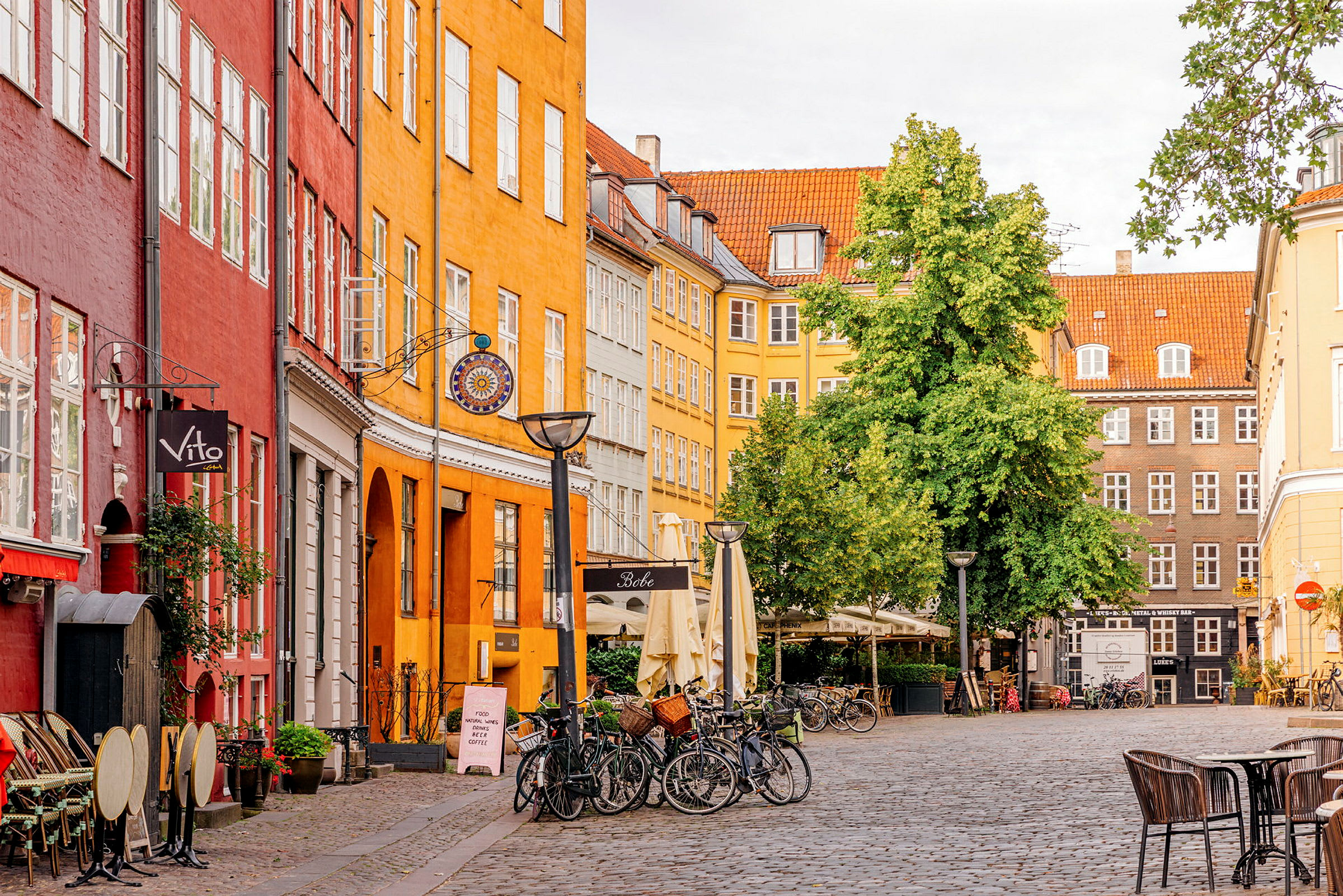 En malerisk brostensgade med farverige bygninger, cykler og udendørs servering under parasoller. (AI-genereret tekst)