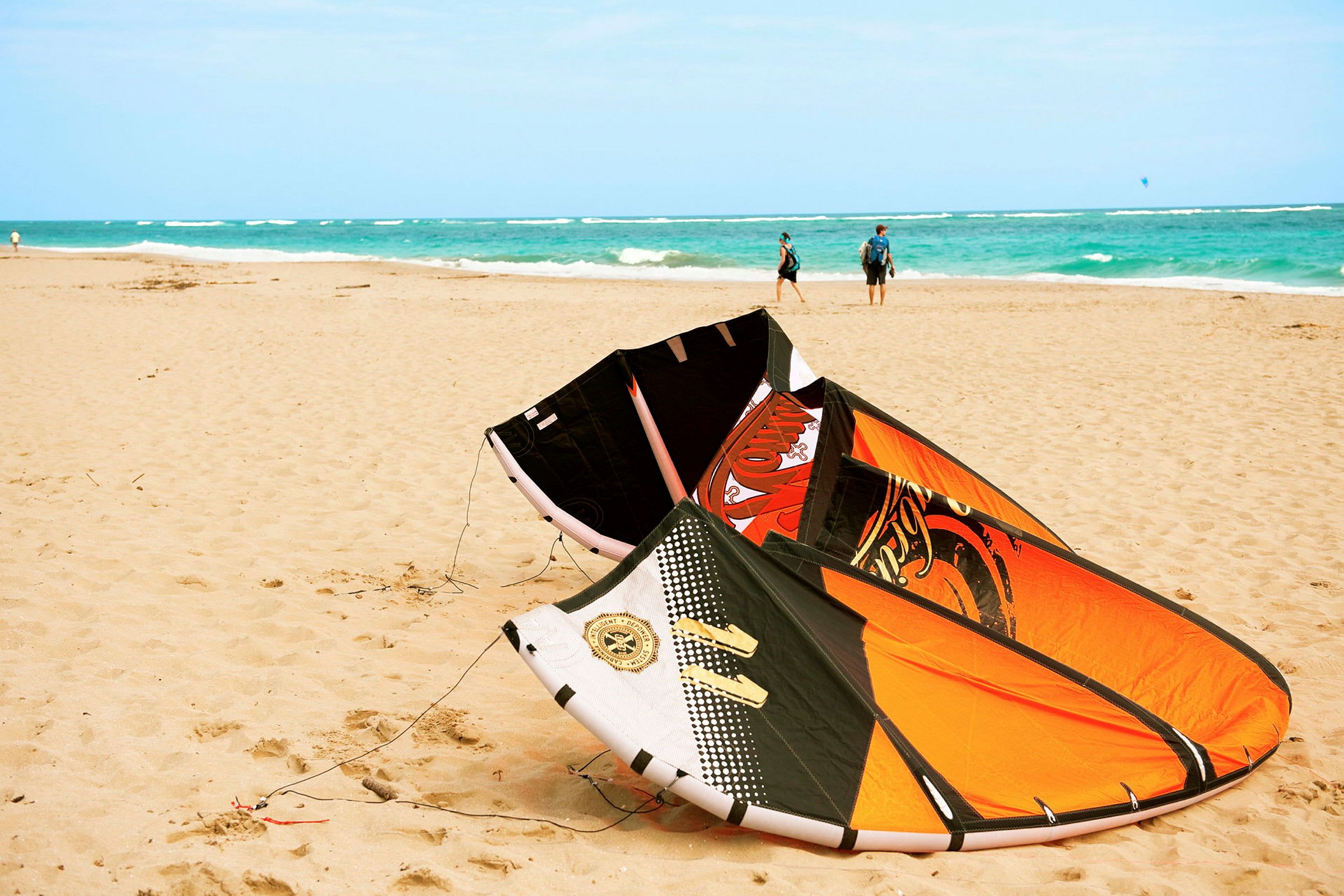 Et strandlandskab med en oppustet kitesurfvinge på sanden, havet i baggrunden og nogle personer gående på stranden. (AI-genereret tekst)