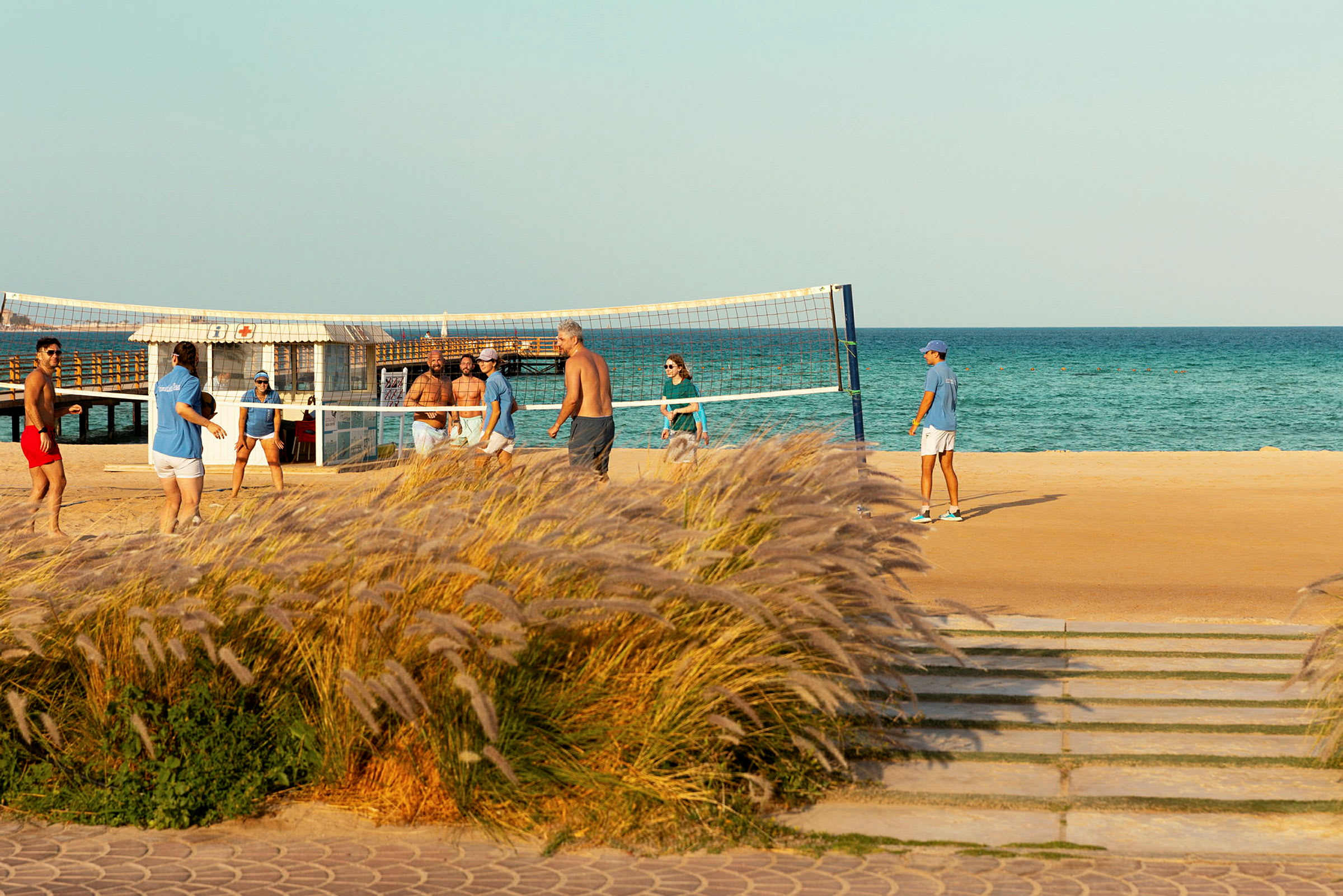 Personer spiller strandvolleyball ved havet, med græs i forgrunden. (AI-genereret tekst)