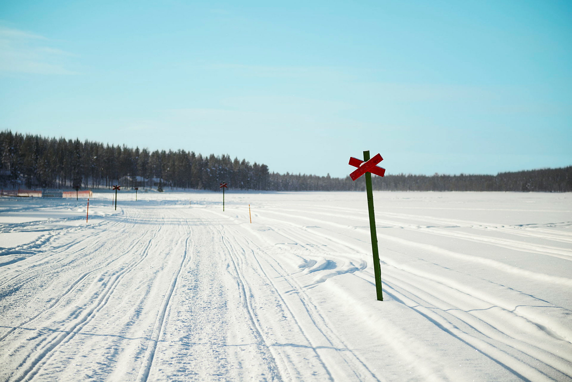 En sneklædt vej med røde kryds markerede vejskilte og skov i baggrunden under en klar blå himmel. (AI-genereret tekst)