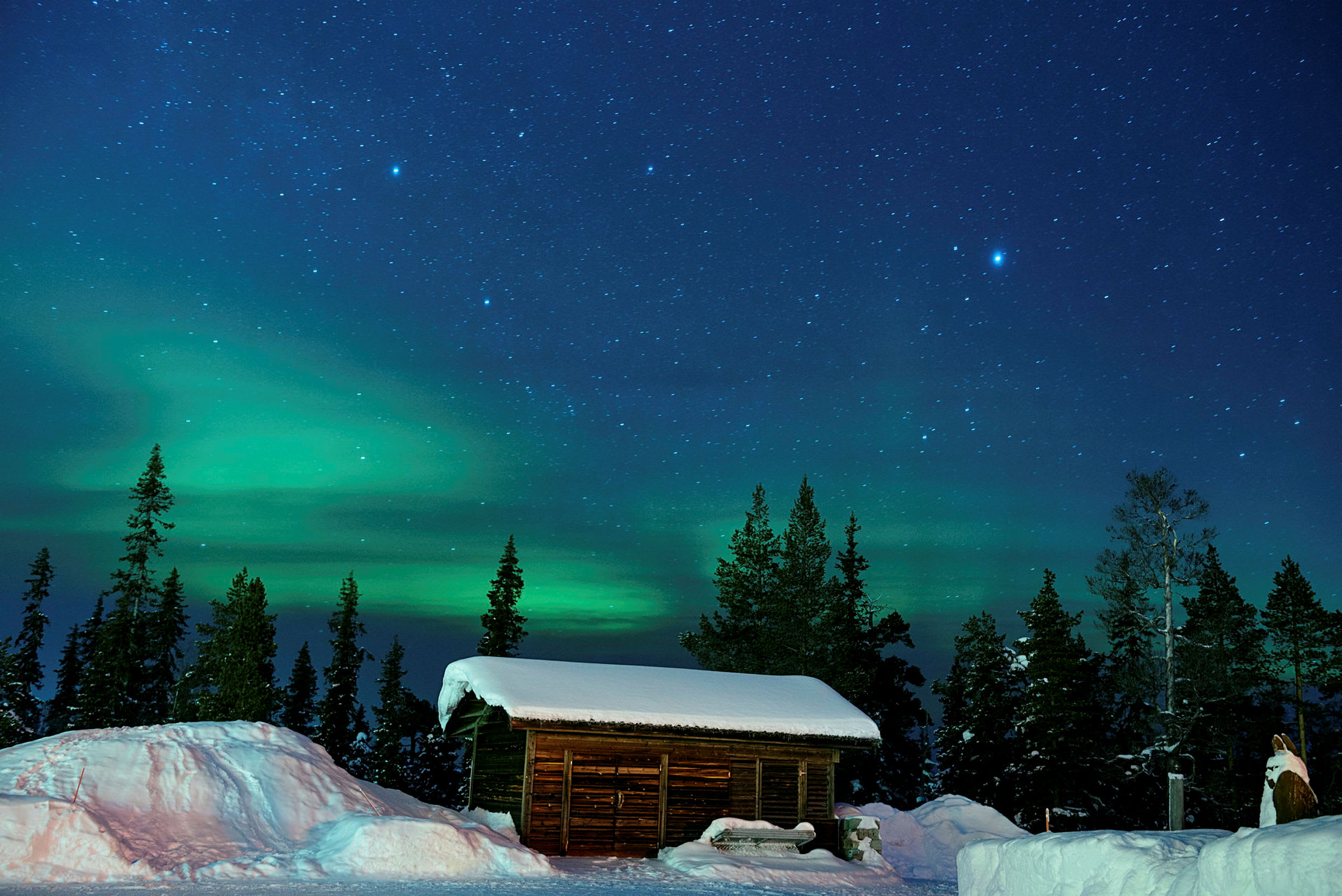 En træhytte med sneklædt tag foran en skov, med stjernehimmel og nordlys i baggrunden. (AI-genereret tekst)