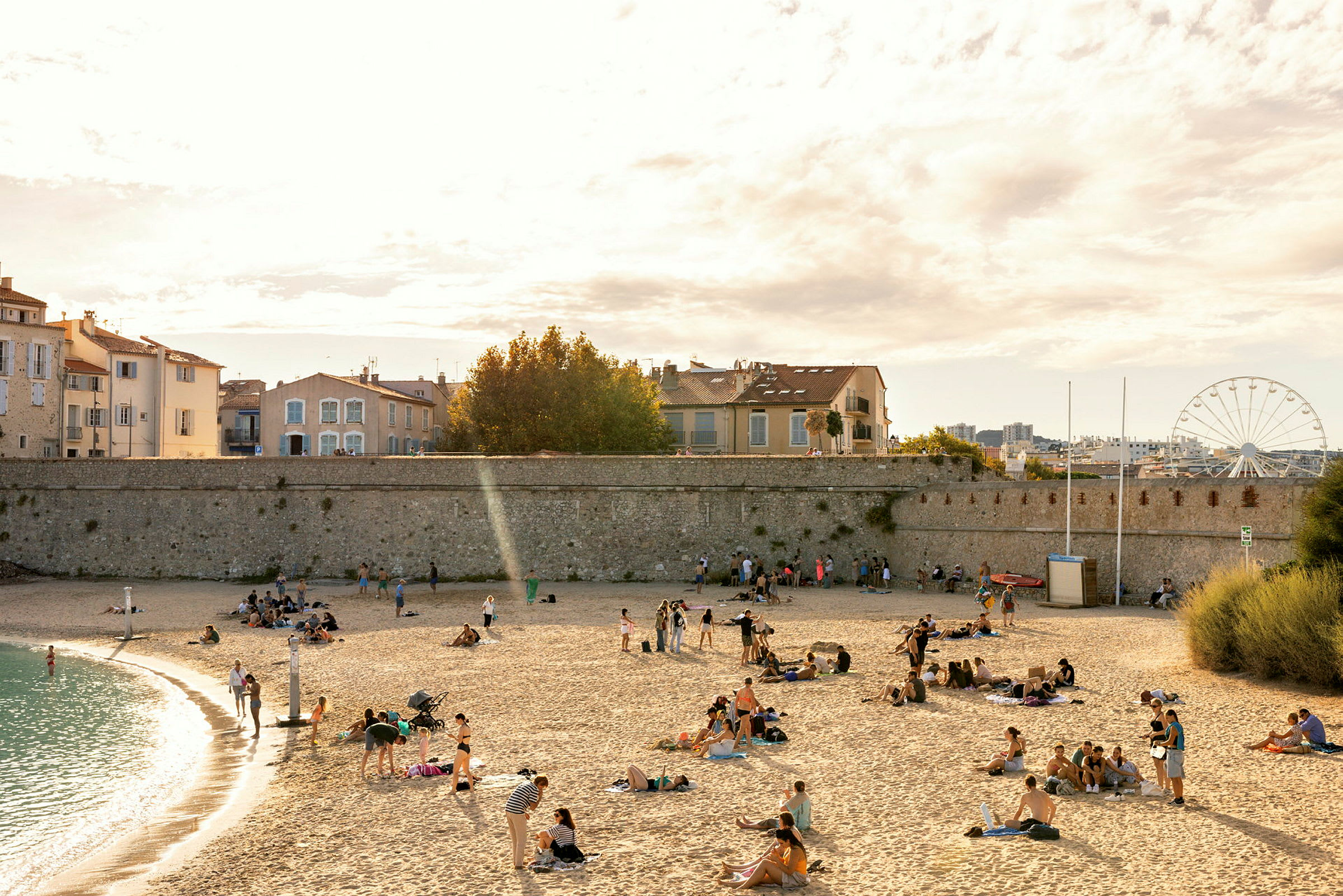 En strand med mennesker, der slapper af på sandet, mens nogle personer spadserer ved vandkanten. Huse og en mur kan ses i baggrunden, samt et pariserhjul. (AI-genereret tekst)