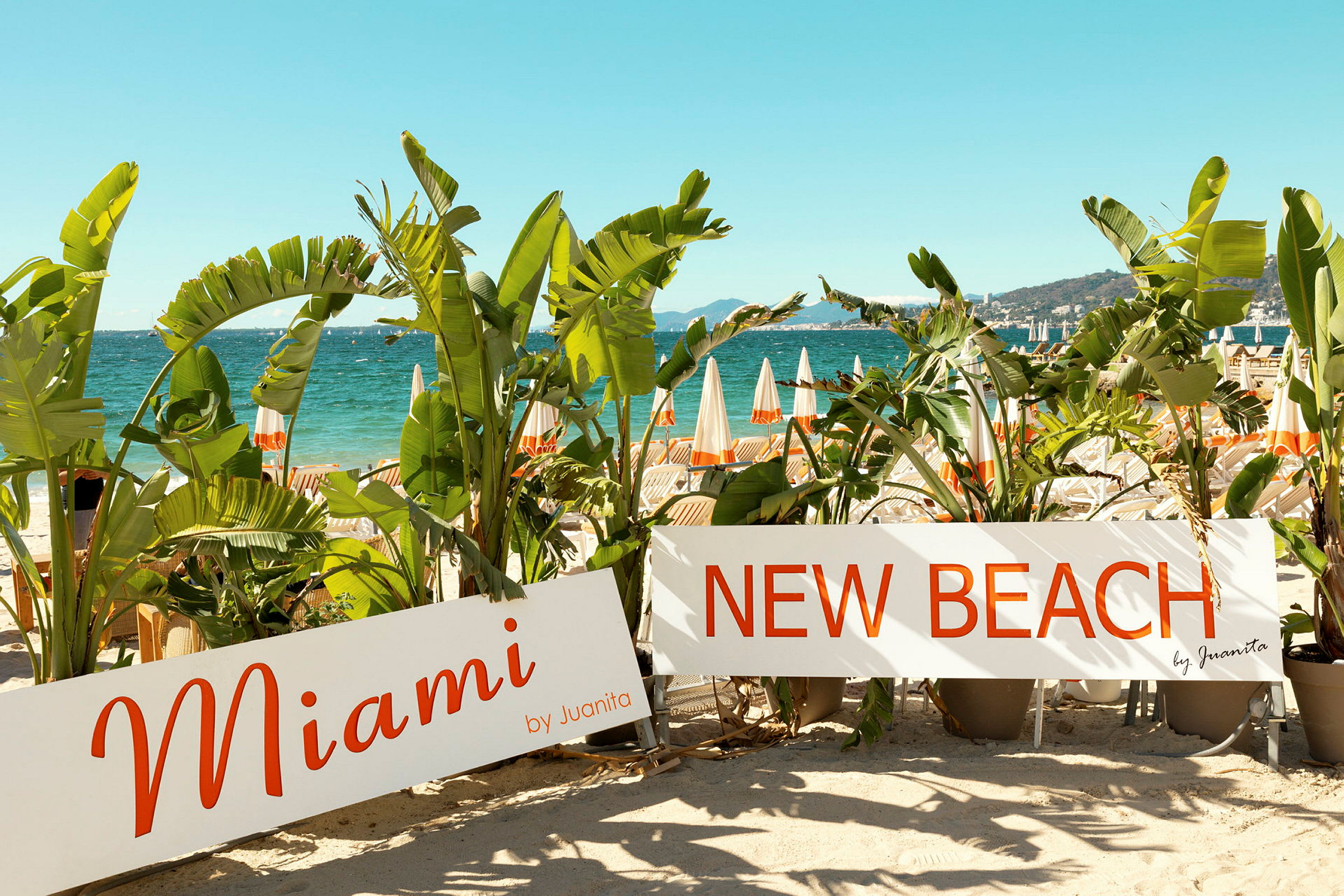 Stranden med parasoller i baggrunden samt palmer og skilte med teksten "Miami" og "New Beach". (AI-genereret tekst)