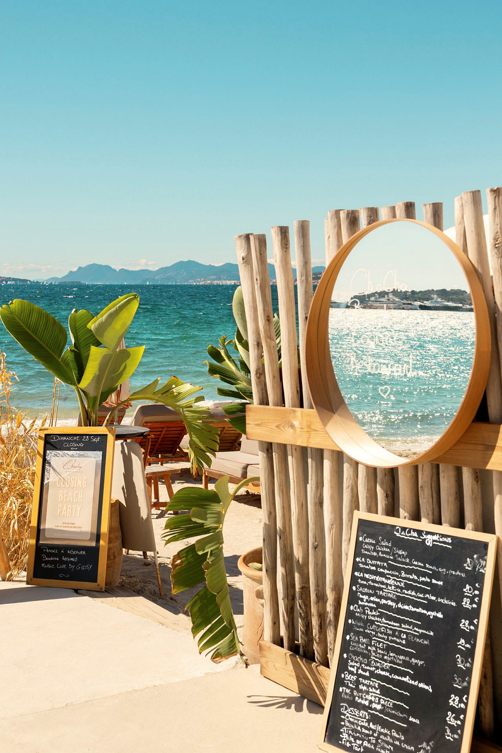 En strand med en træramme og et spejl, omgivet af tropiske planter. En menu står på jorden ved siden af et skilt. (AI-genereret tekst)