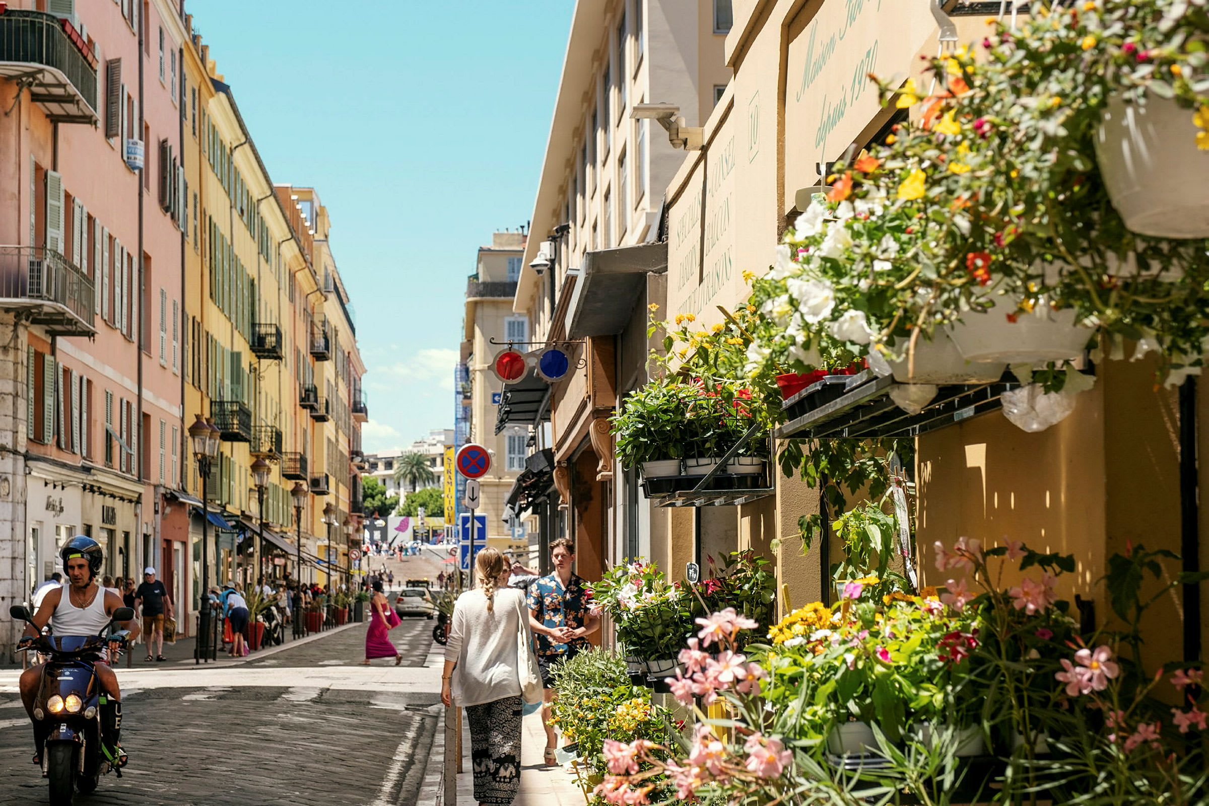 En byvej med stribede farverige bygninger. Blomster hænger på facaden, og folk går og kører på scooter på vejen. (AI-genereret tekst)