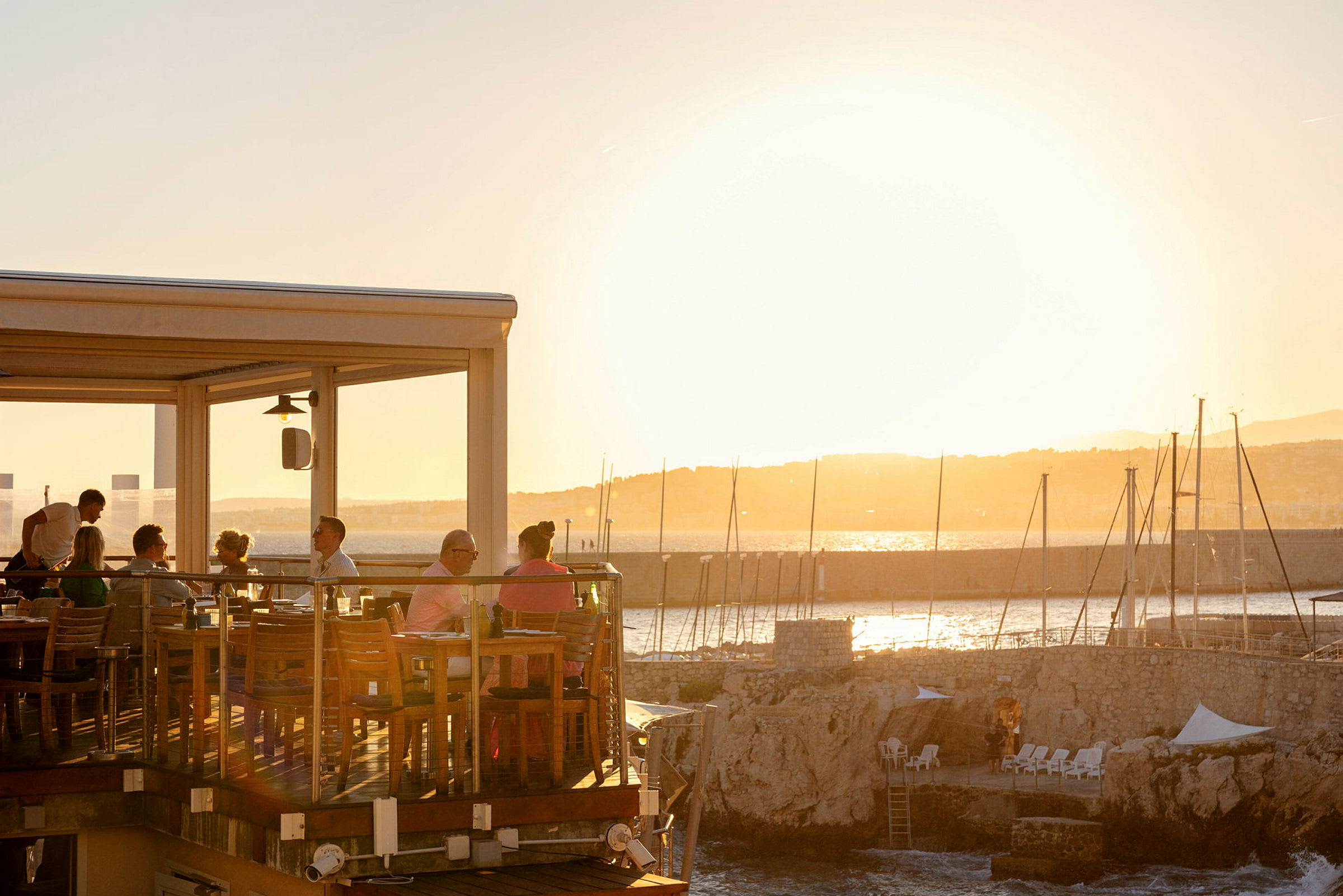 En udendørs terrasse på en restaurant ved havet, hvor gæster sidder og nyder solnedgangen. (AI-genereret tekst)