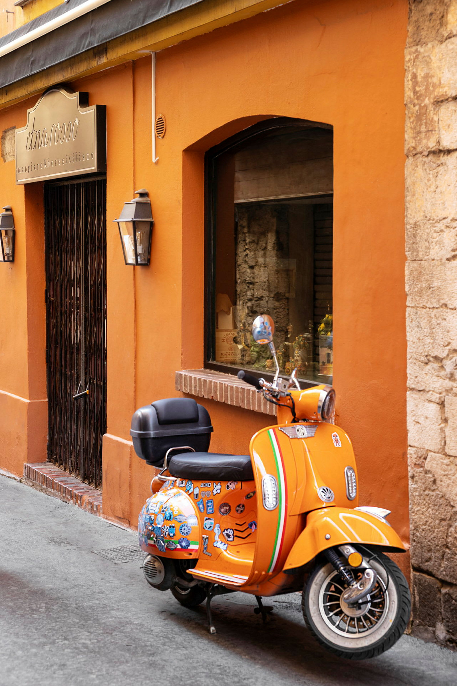 En orange scooter med dekorative klistermærker står parkeret ved en orange bygning med en smedejernsport og et skiltet vindue. (AI-genereret tekst)