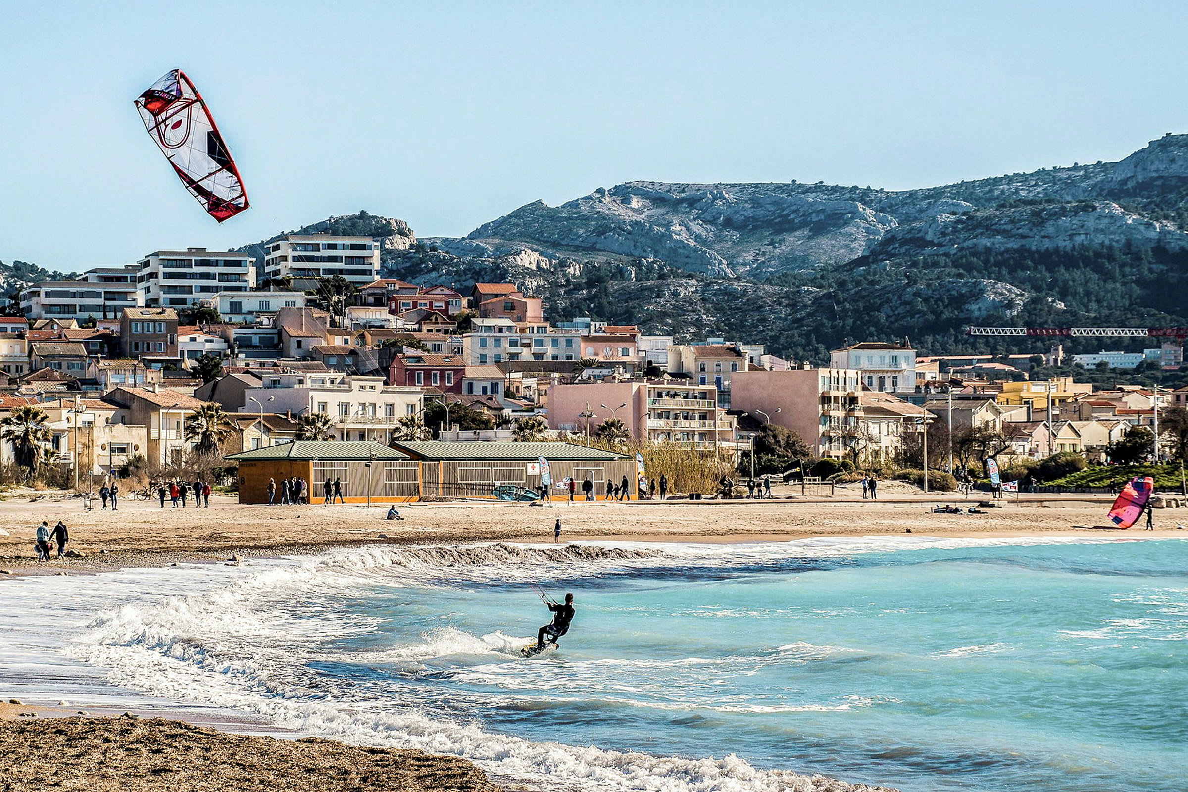 Strand med kitesurfere og bølger, huse synlige i baggrunden. (AI-genereret tekst)
