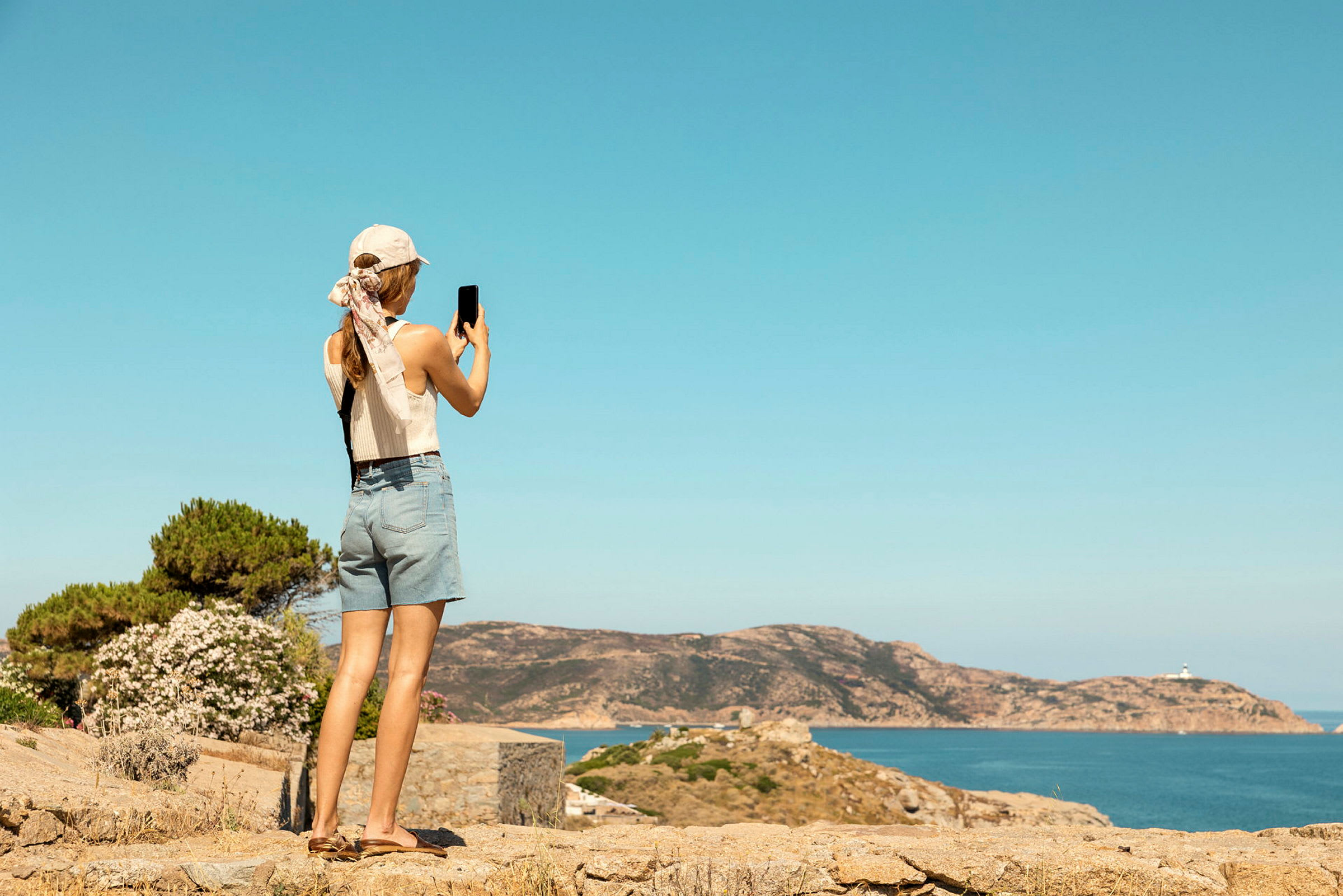 En person med hat og shorts tager et billede af en kystudsigt med hav og klipper. (AI-genereret tekst)