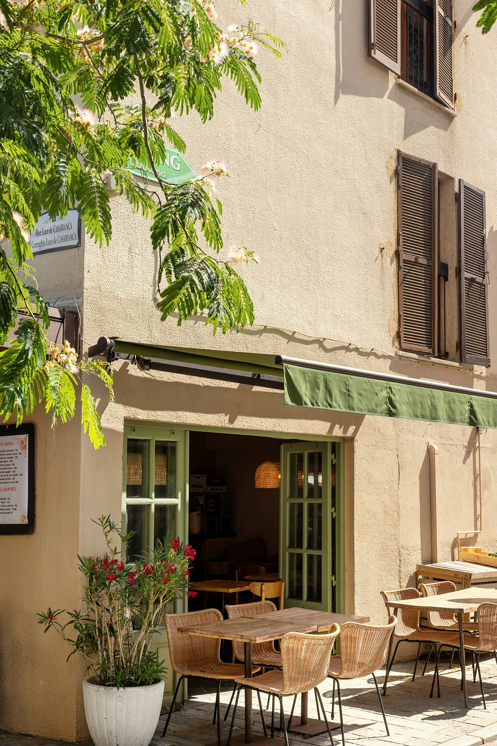 En café med udendørs servering, hvor borde og stole står på en brolagt gårdsplads. Grønt områder og blomster omgiver bygningen. (AI-genereret tekst)