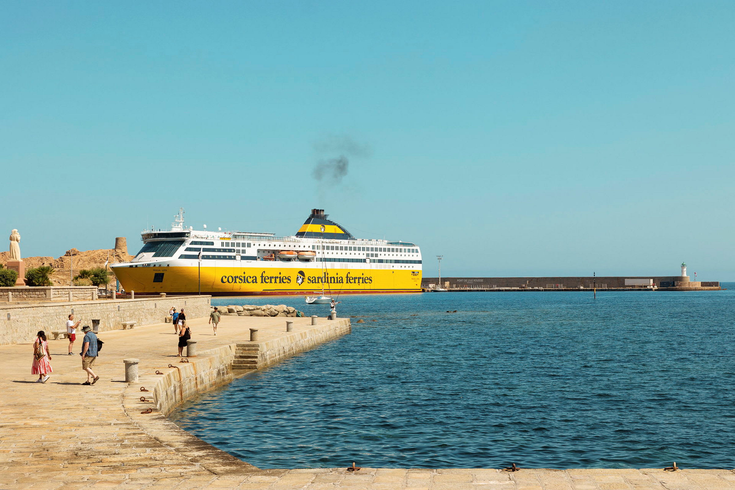 En gul og hvid færge fra Corsica Ferries ligger ved kajen, med mennesker der går på kajområdet og havet synligt i forgrunden. (AI-genereret tekst)