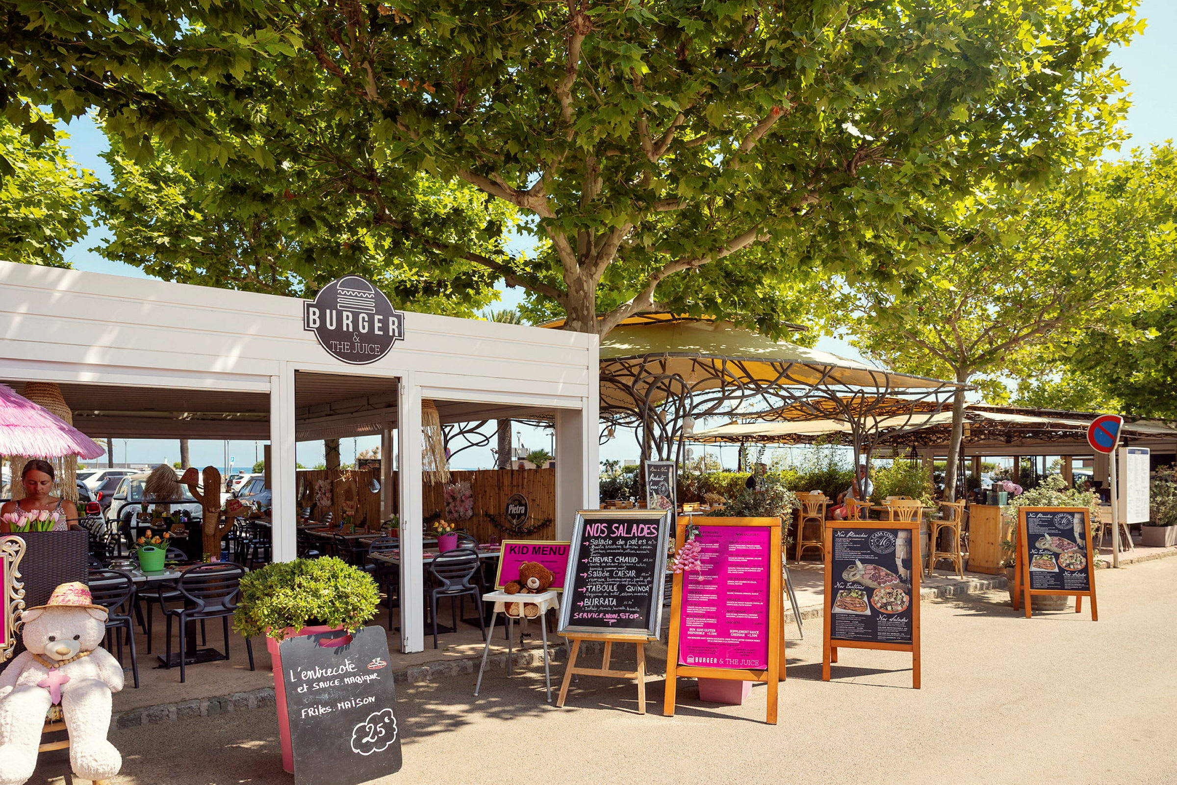 Udendørs restaurant med skilte og beplantning. Borde og stole under trækroner og parasoller. (AI-genereret tekst)