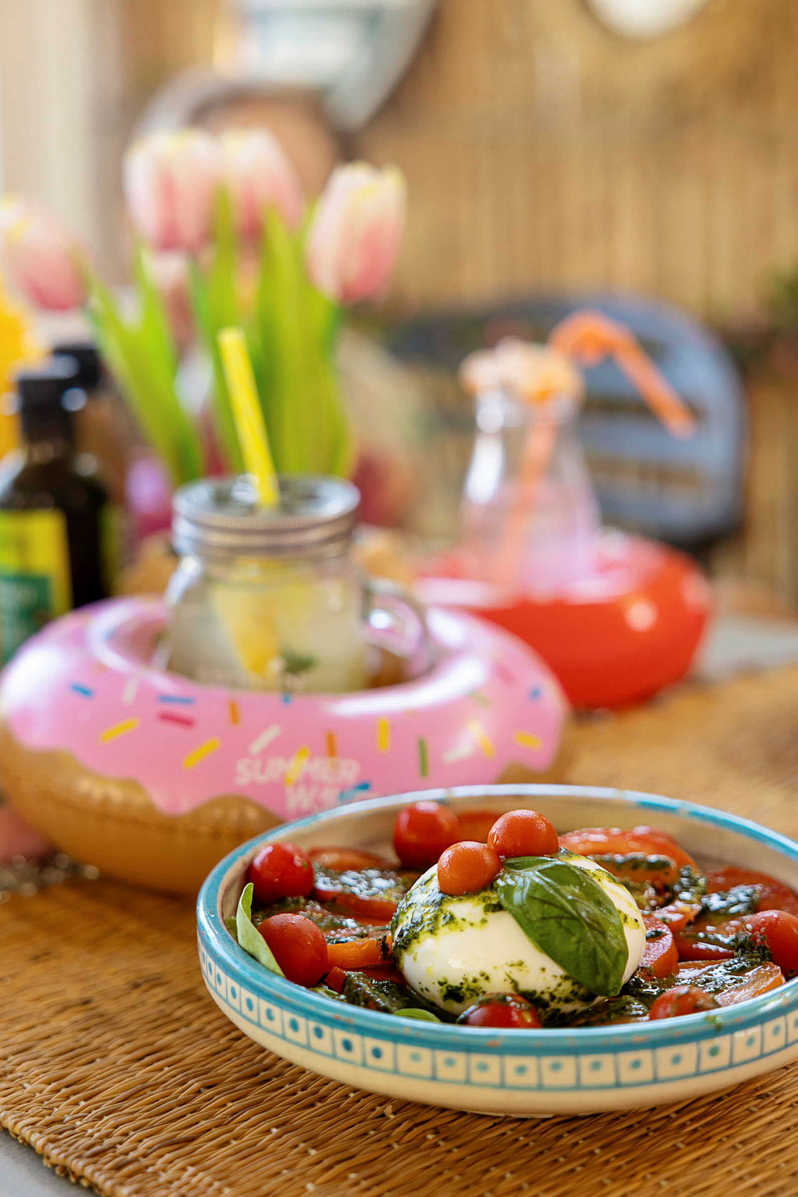 En ret med buffalo-mozzarella, tomater og basilikum samt drikkevarer med dekorationer. Baggrunden viser tulipaner og andre pyntegenstande. (AI-genereret tekst)