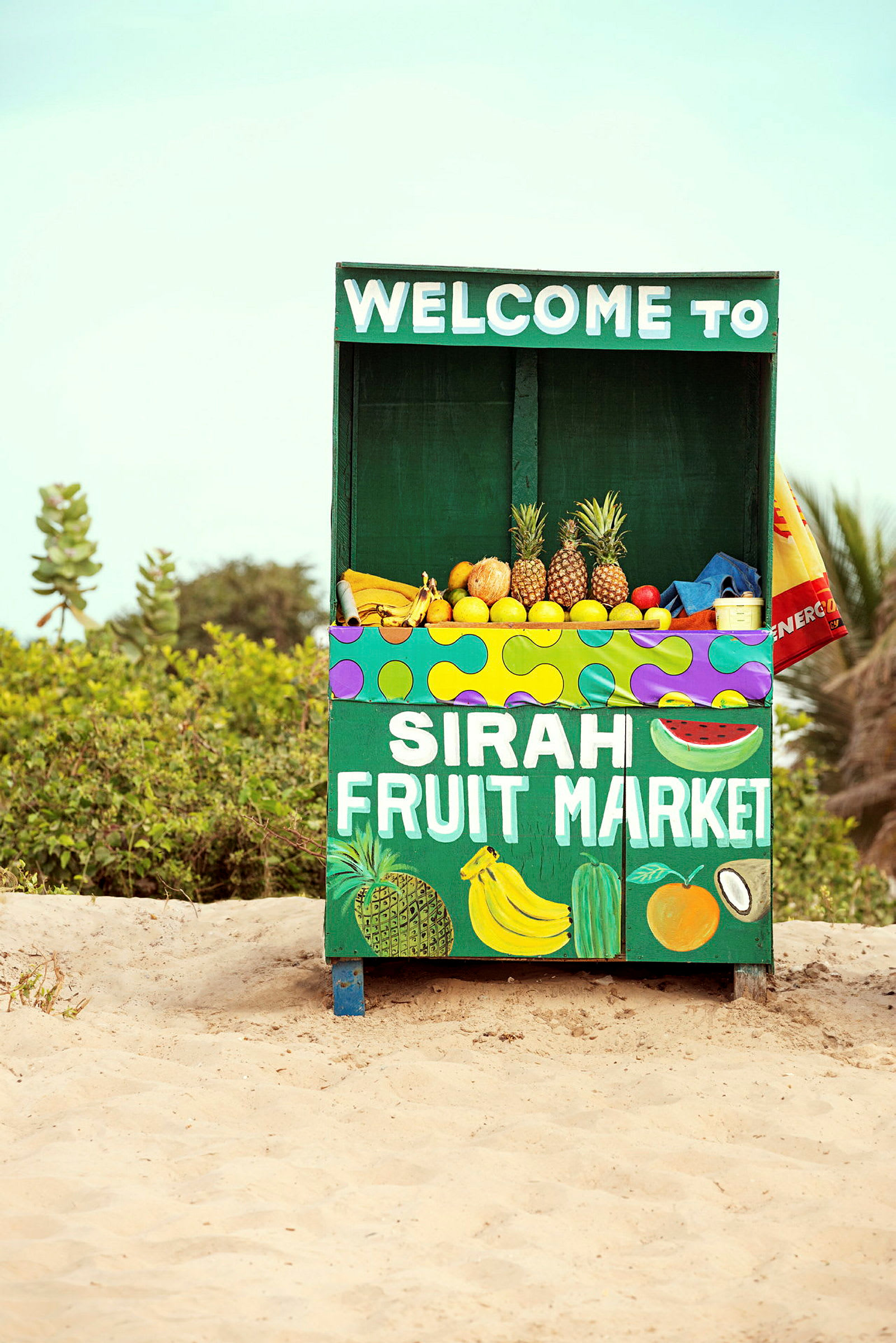 Et frugtstativ med teksten "Sirah Fruit Market" tilbyder forskellige frugter, herunder ananas, bananer og citrusfrugter. Stativet står på sand med vegetation i baggrunden. (AI-genereret tekst)