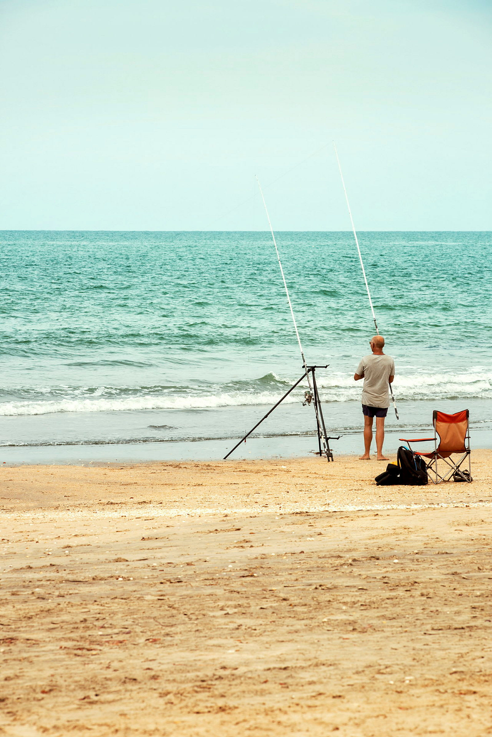 En person står ved strandkanten med to fiskestænger på et stativ. En stol står i sanden ved siden af. (AI-genereret tekst)