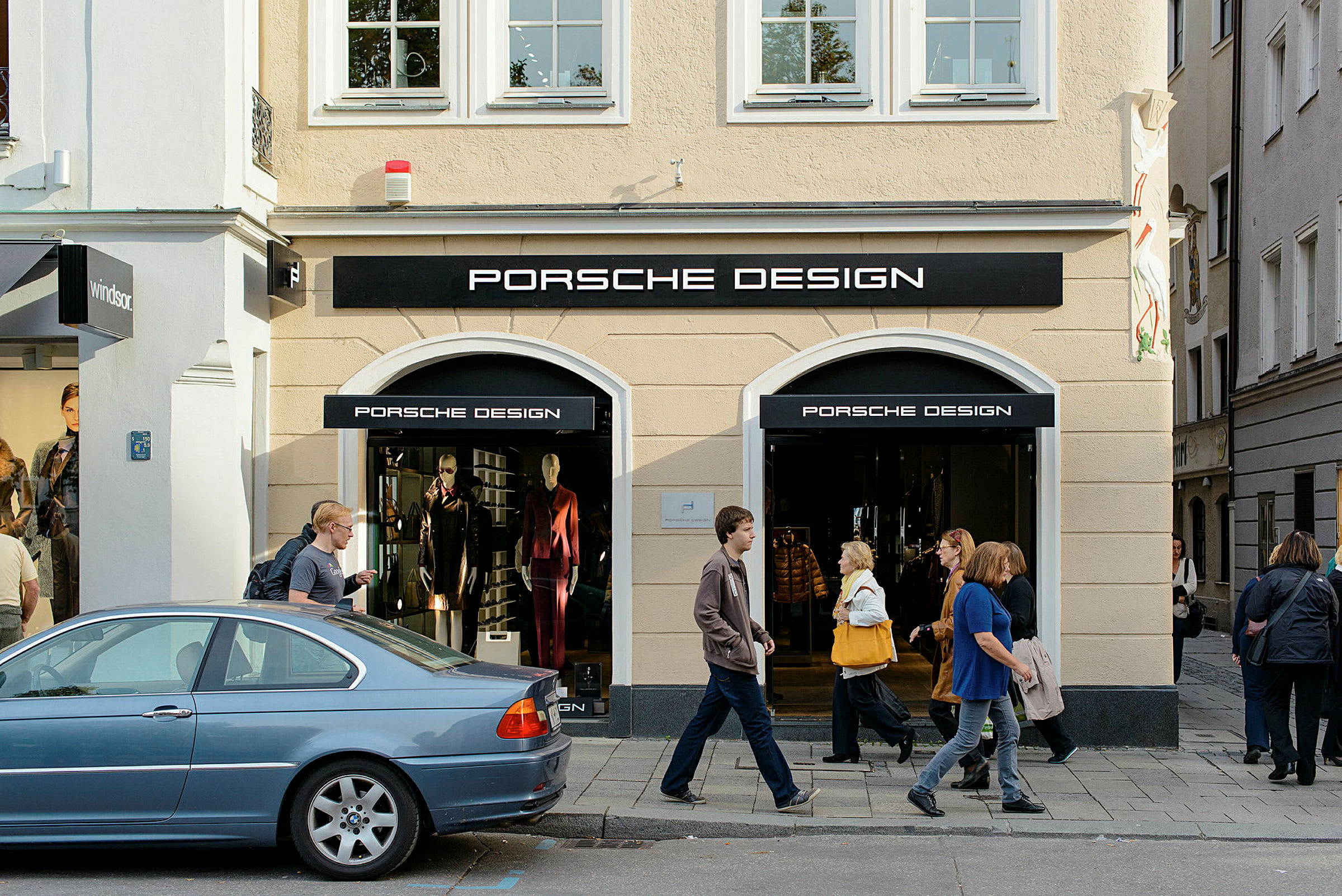 Indgang til en butik med Porsche Design-logo, med udstillingsvinduer og mennesker, der går på fortovet udenfor. (AI-genereret tekst)