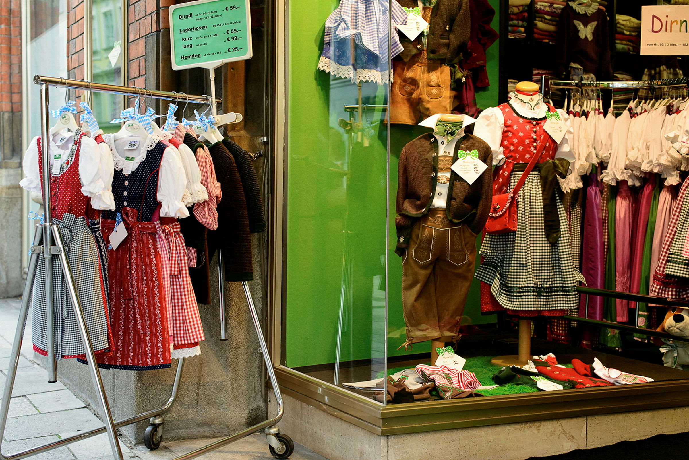 Et butikvindue med traditionelle folkedragter samt et tøjstativ udenfor fyldt med dirndl-kjoler. (AI-genereret tekst)