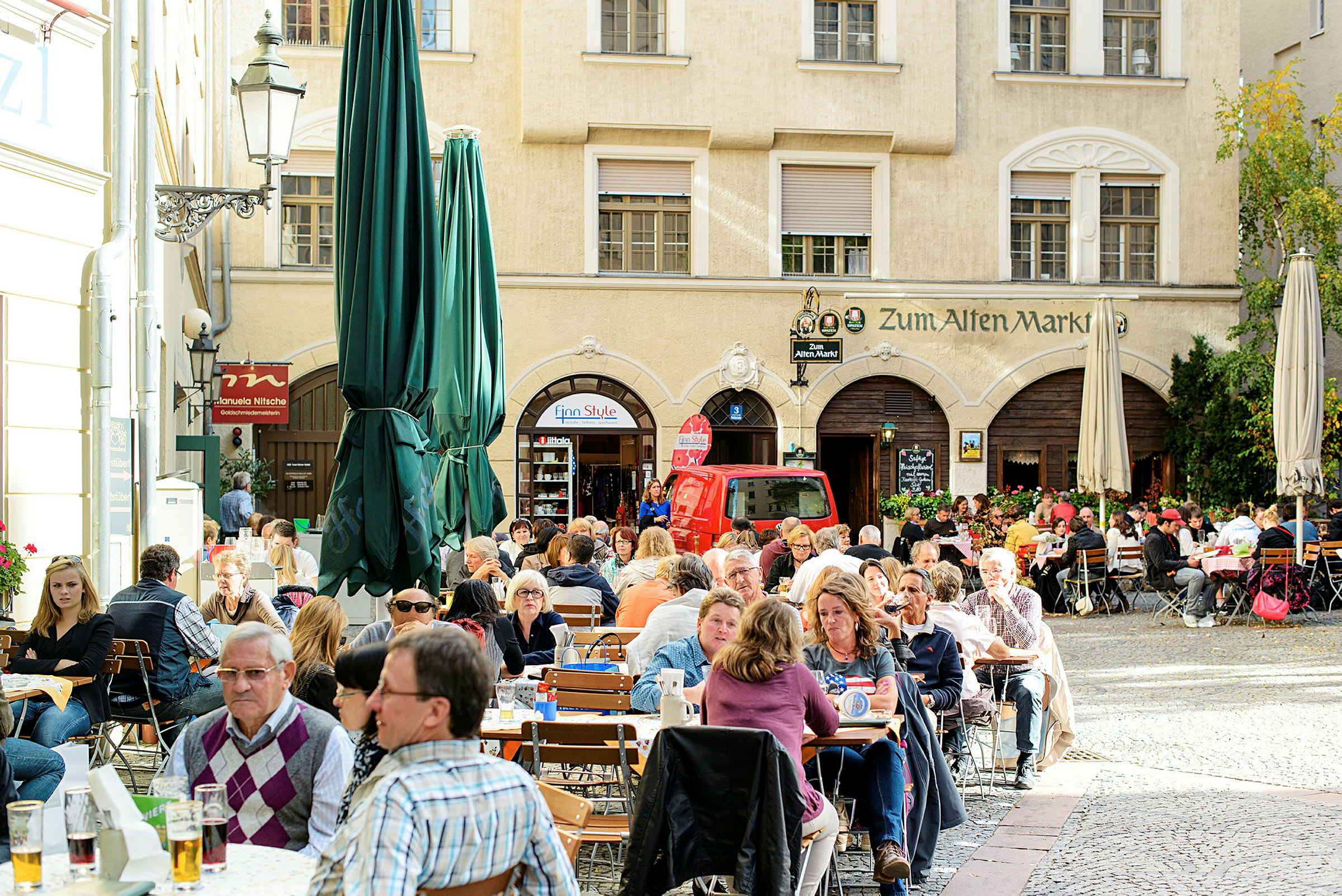 Udendørs servering med mange mennesker, der sidder ved træborde på en brostensbelagt terrasse omgivet af gamle bygninger. (AI-genereret tekst)