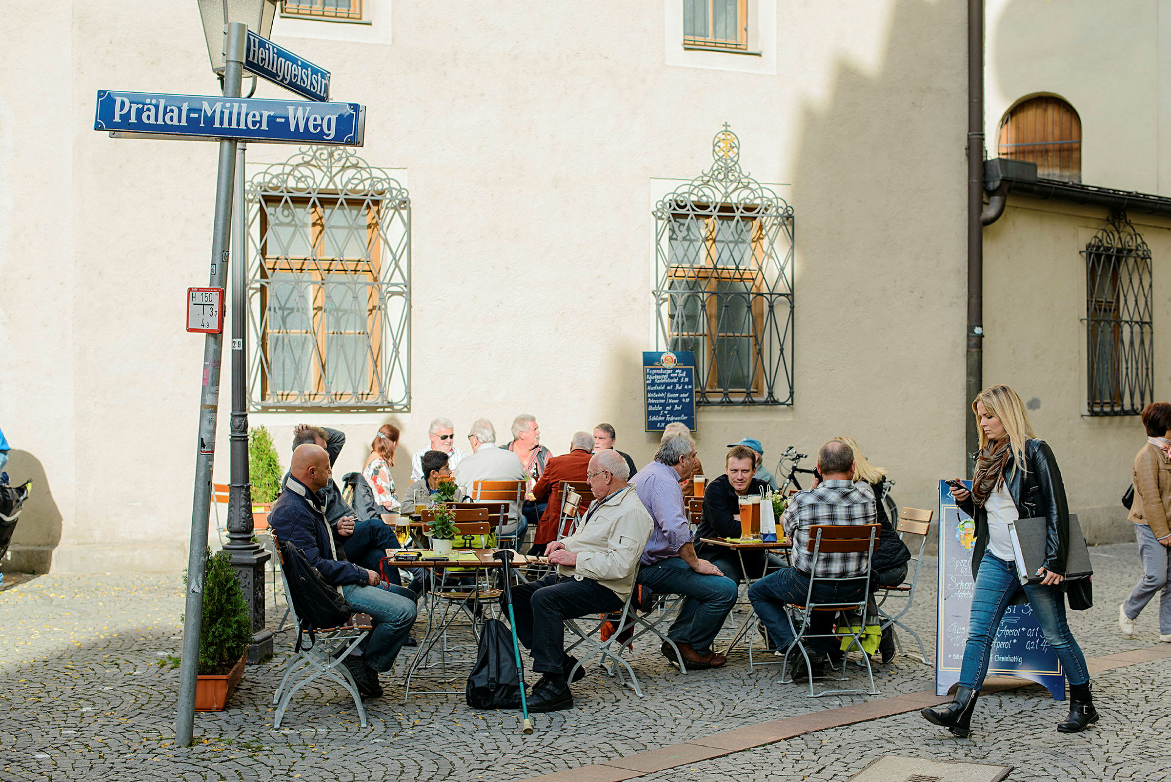Folk nyder mad og drikke ved caféborde udendørs på en brostensgade. (AI-genereret tekst)