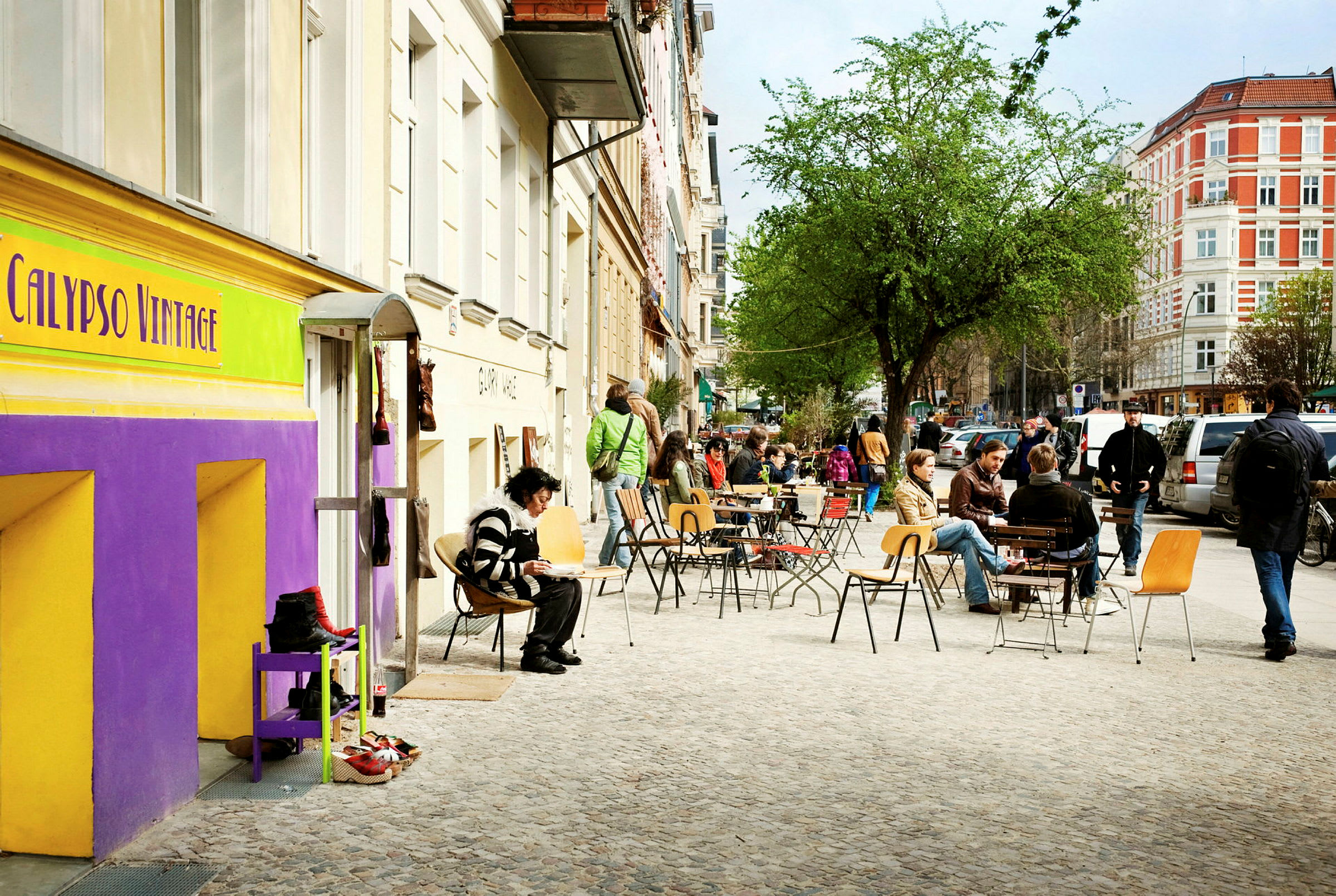 Gaden er omgivet af ældre bygninger med caféborde og udendørs siddepladser, hvor folk sidder og nyder dagen. (AI-genereret tekst)