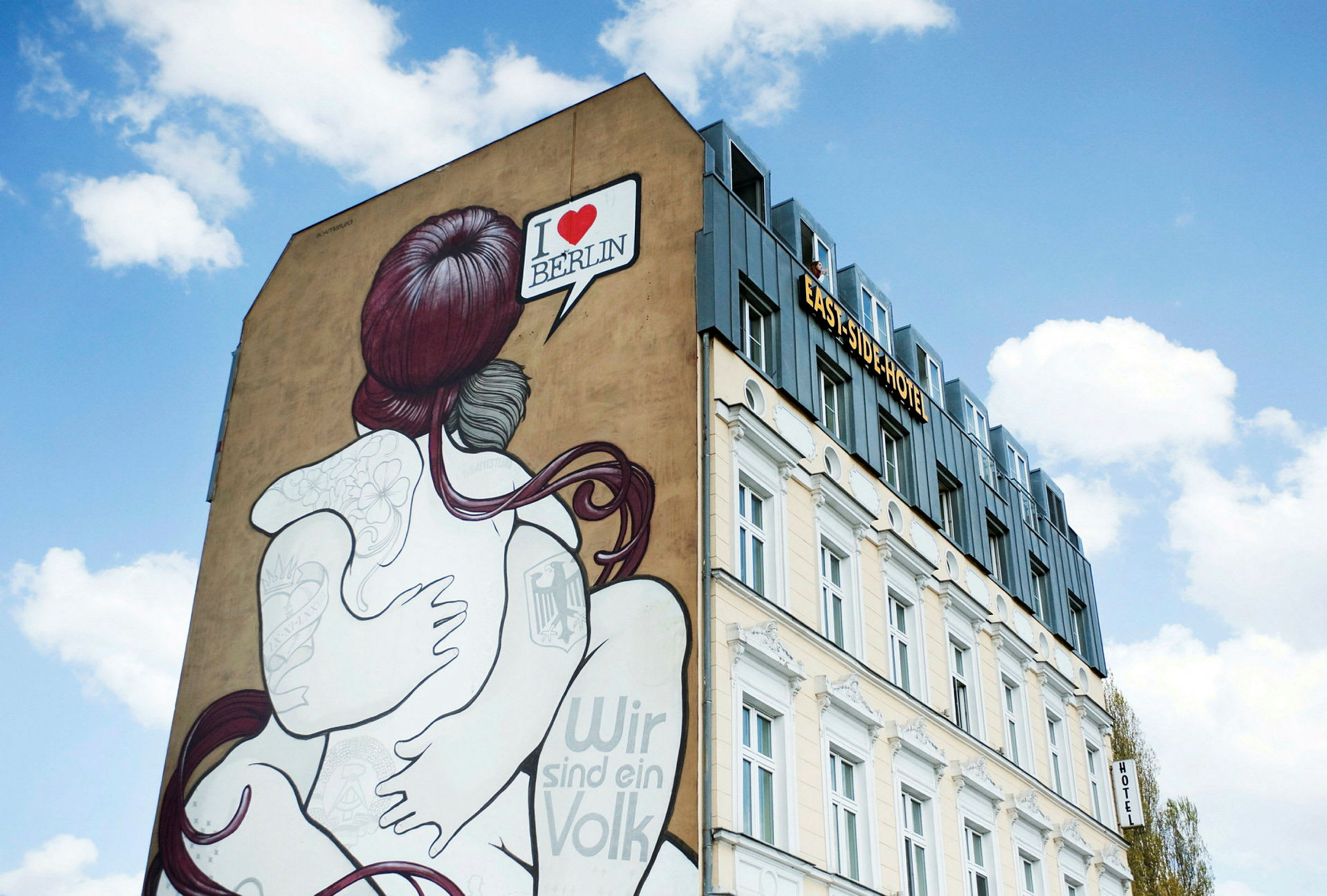 En bygning med et vægmaleri af et par, der omfavner hinanden. Teksten "I ♥ BERLIN" vises i en taleboble. Himlen er klar med hvide skyer. (AI-genereret tekst)