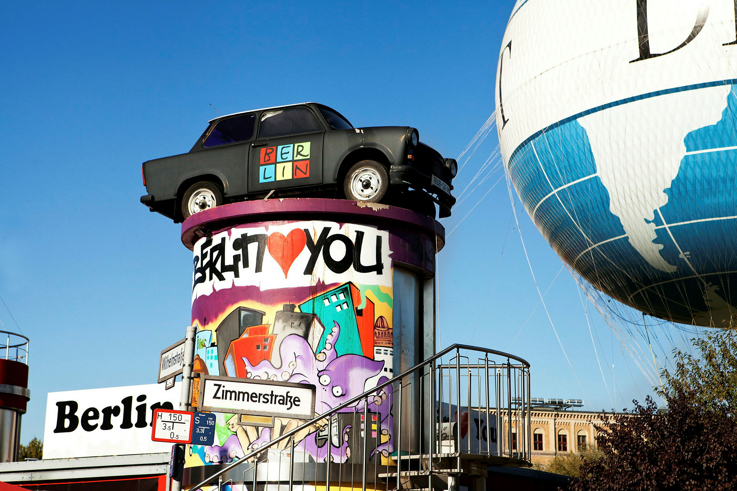 Et billede af en sort bil placeret på toppen af en farverigt graffiti-dekoreret søjle ved siden af en luftballon og et skilt med teksten Zimmerstraße. (AI-genereret tekst)