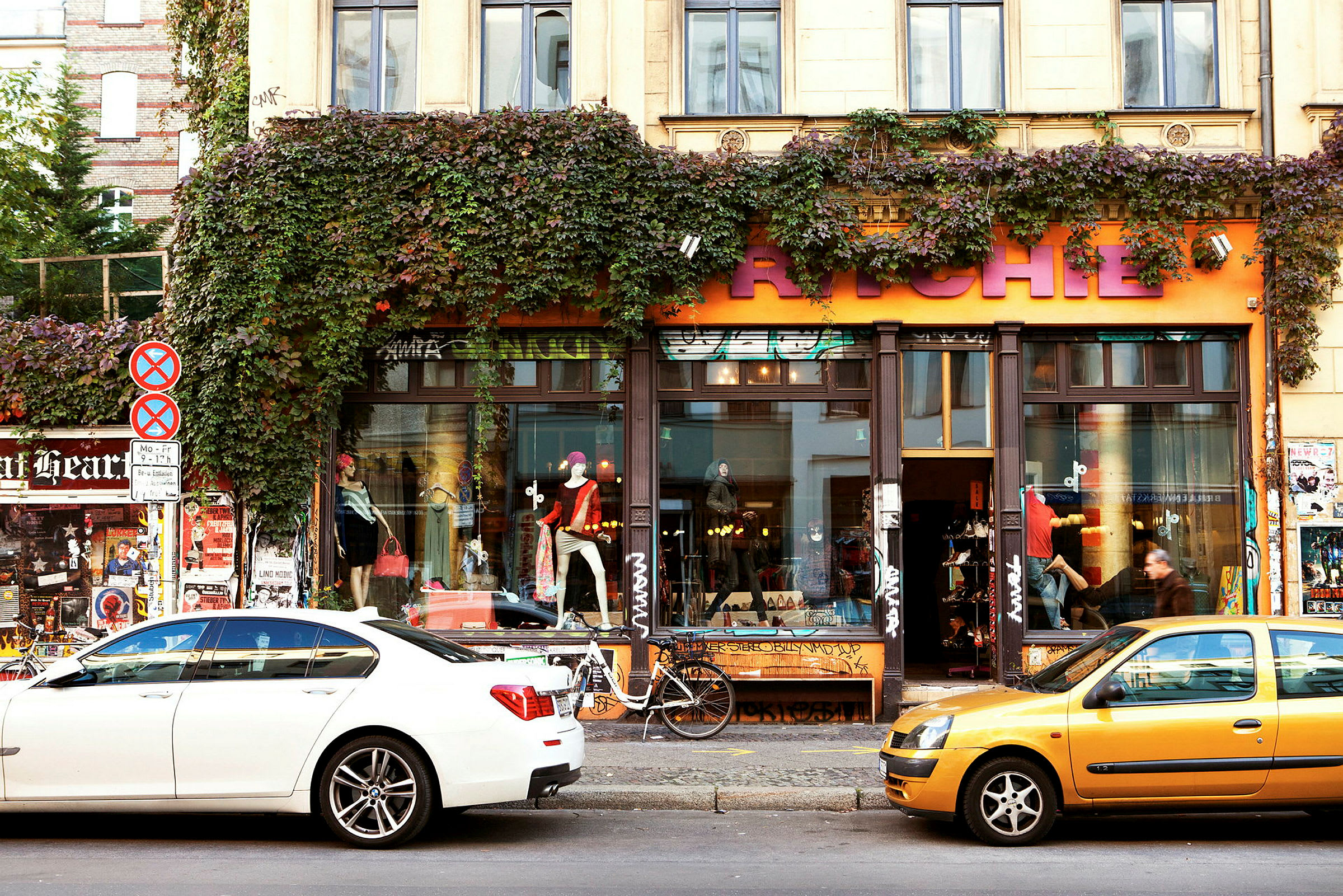 En bygning med en butik har et orange skilt med store bogstaver, omgivet af grønne planter, der klatrer langs facaden. Biler er parkeret foran butikken. (AI-genereret tekst)