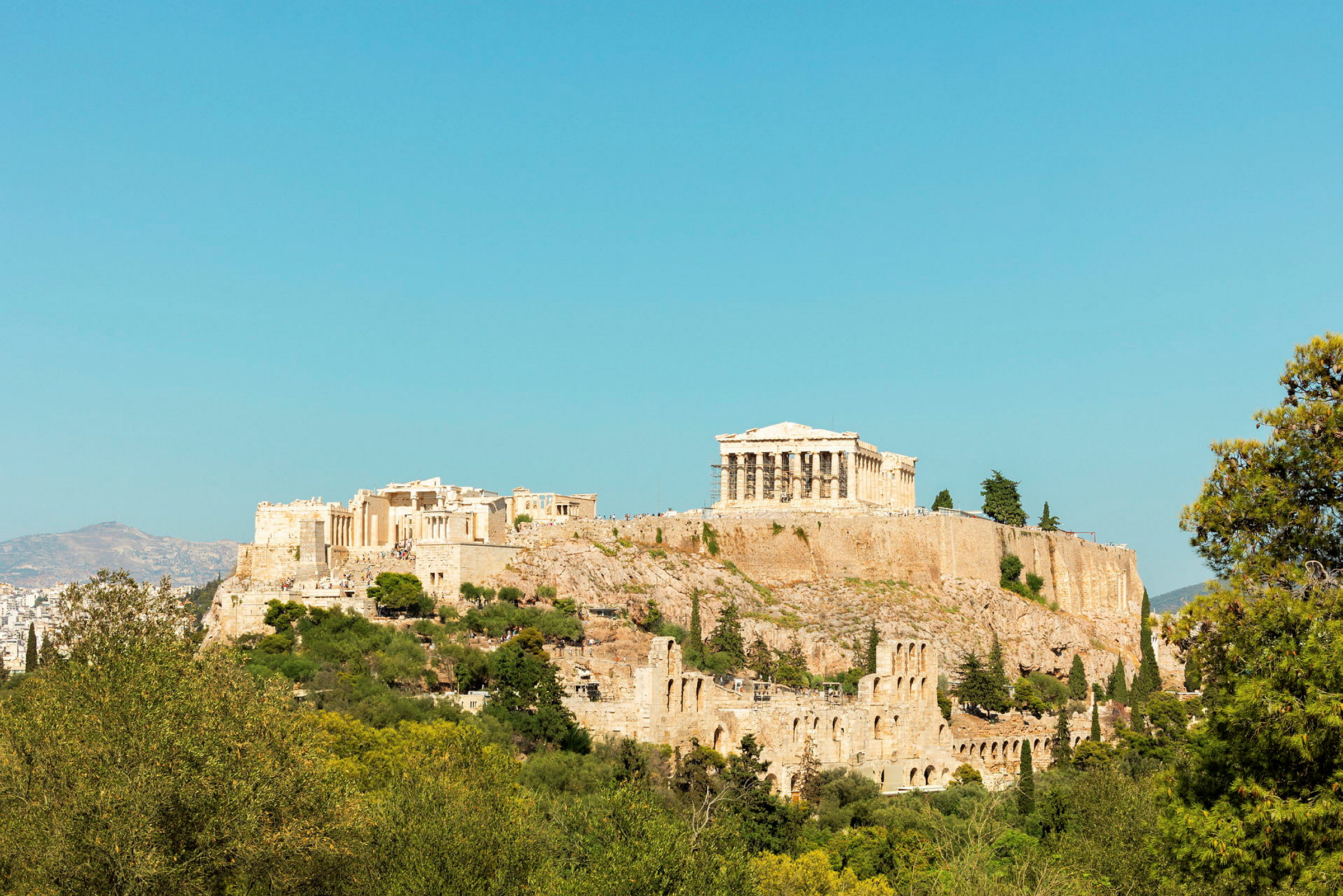 Fotografi af Akropolis med Parthenon på toppen, omgivet af grønt og med klar blå himmel i baggrunden. (AI-genereret tekst)