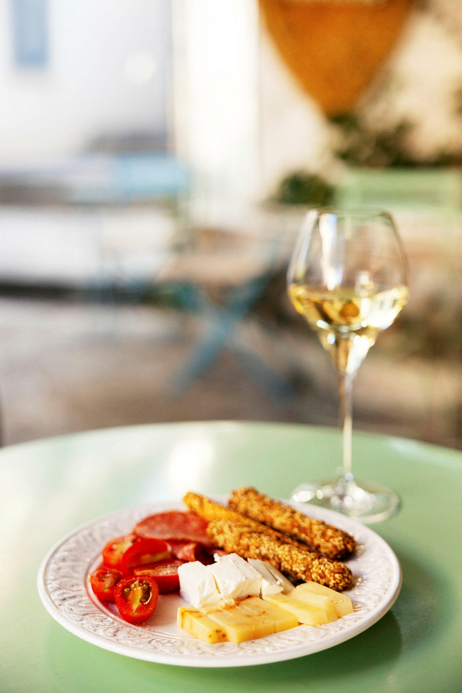 En tallerken med ost, brødstænger og cherrytomater, ved siden af et glas hvidvin, placeret på et grønt bord. (AI-genereret tekst)