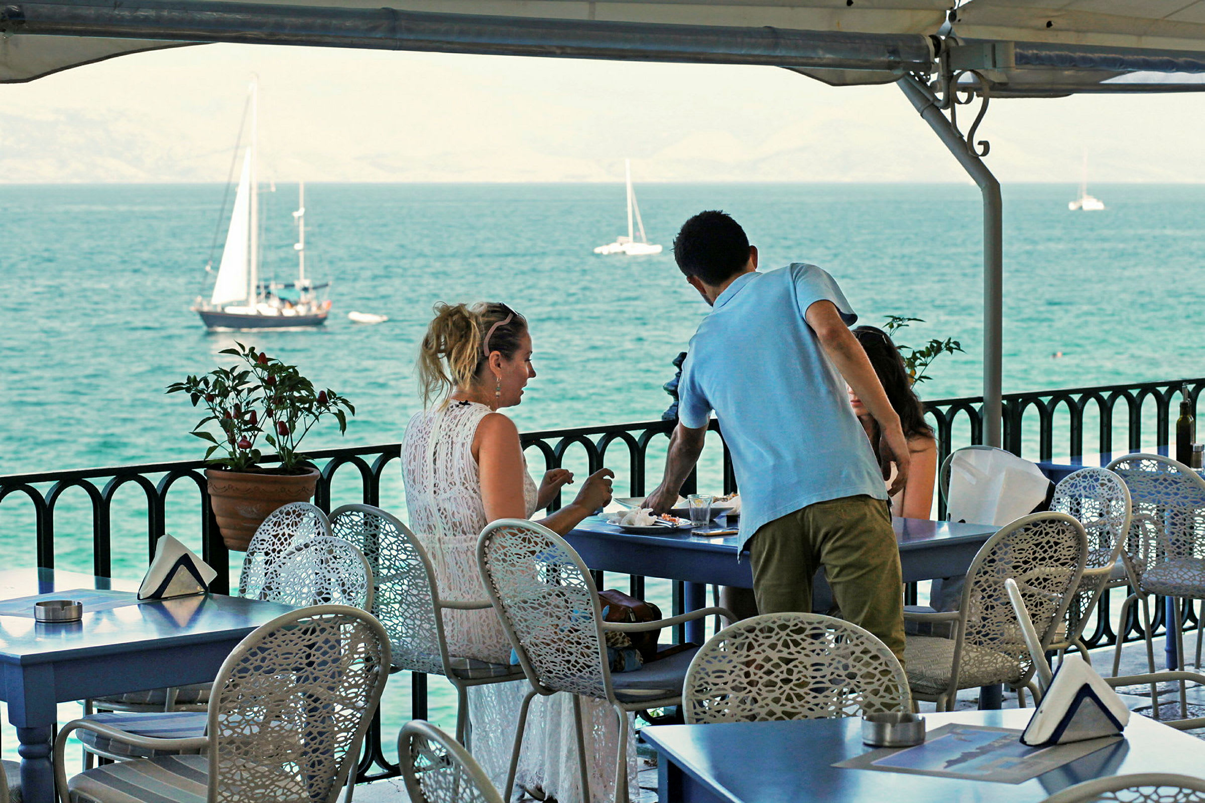 Folk sidder ved et bord på en terrasse med udsigt over havet og sejlbåde. (AI-genereret tekst)