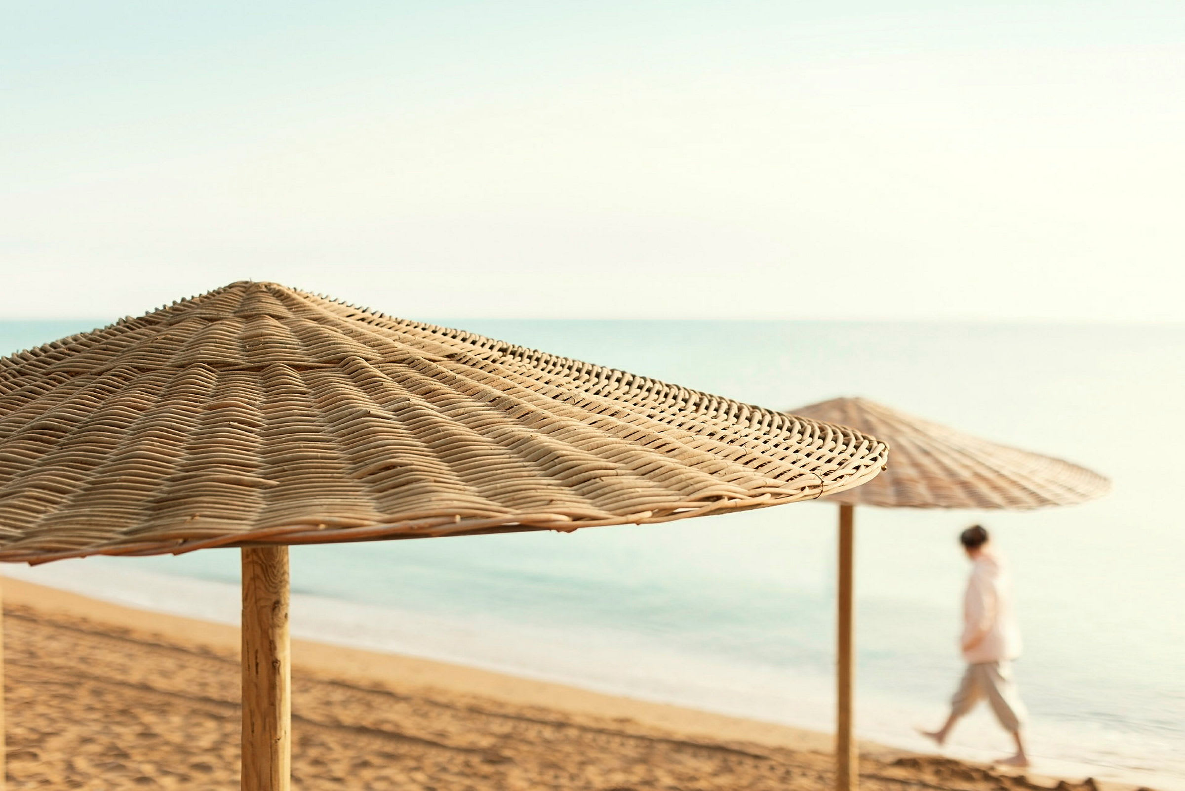 Strand med to solparasoller af rattan og en person, der går ved vandet. (AI-genereret tekst)