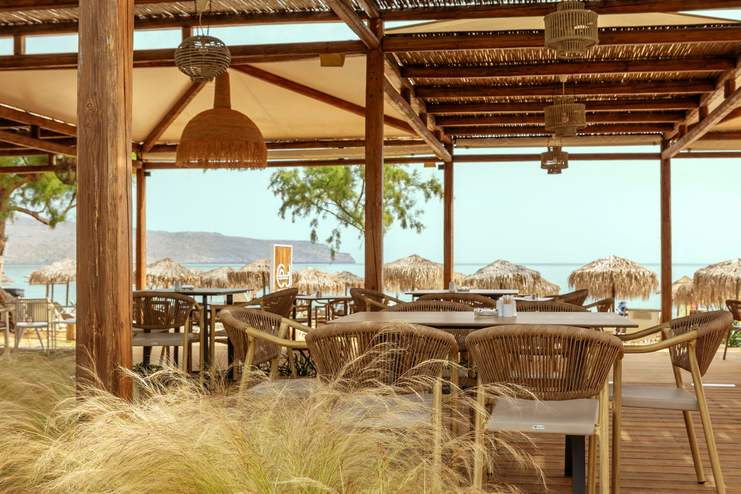 En udendørs restaurant med borde og stole under en trækonstruktion. Baggrunden viser stranden og havet. (AI-genereret tekst)