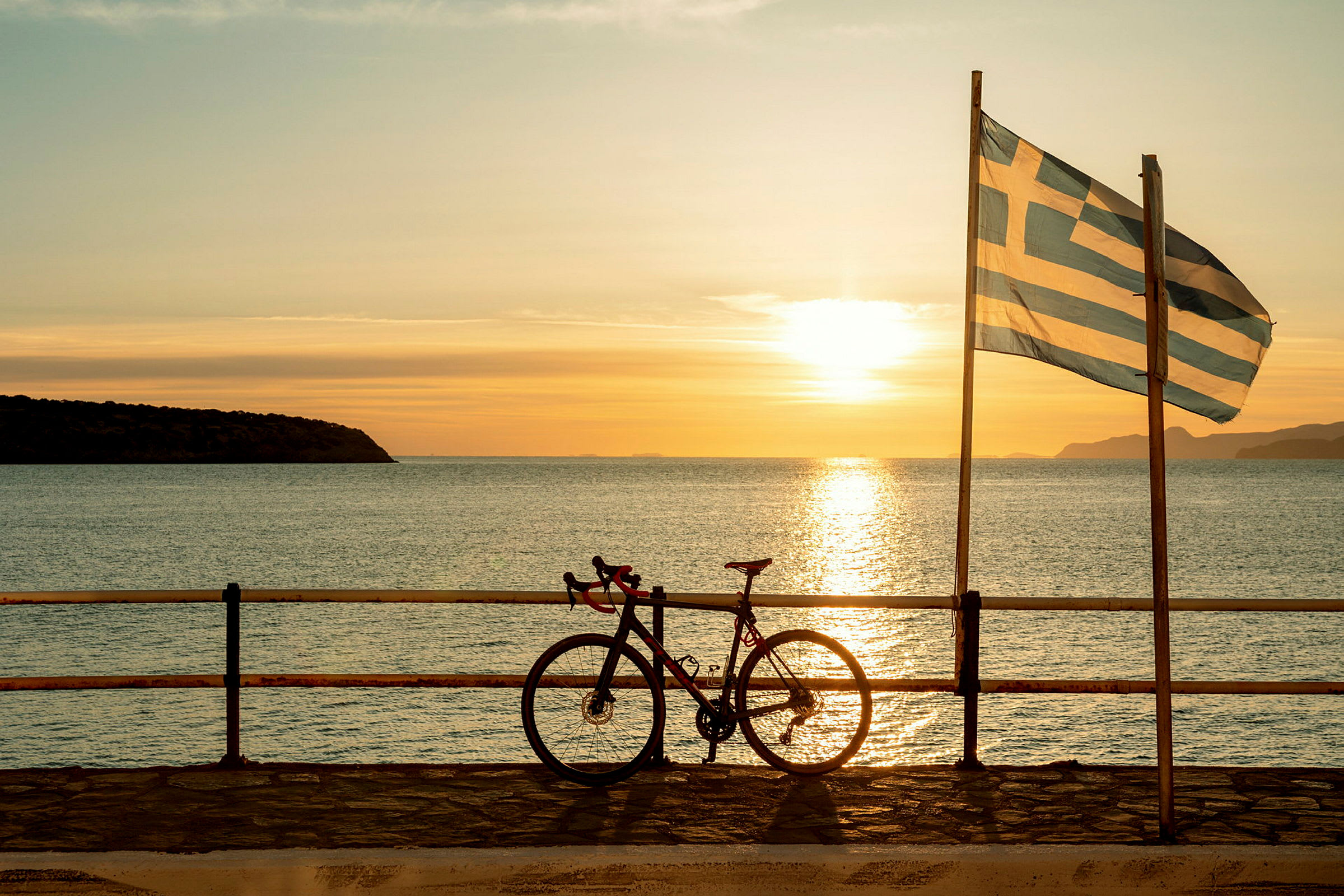 En cykel står op ad et rækværk ved havet med et græsk flag og en solnedgang i baggrunden. (AI-genereret tekst)