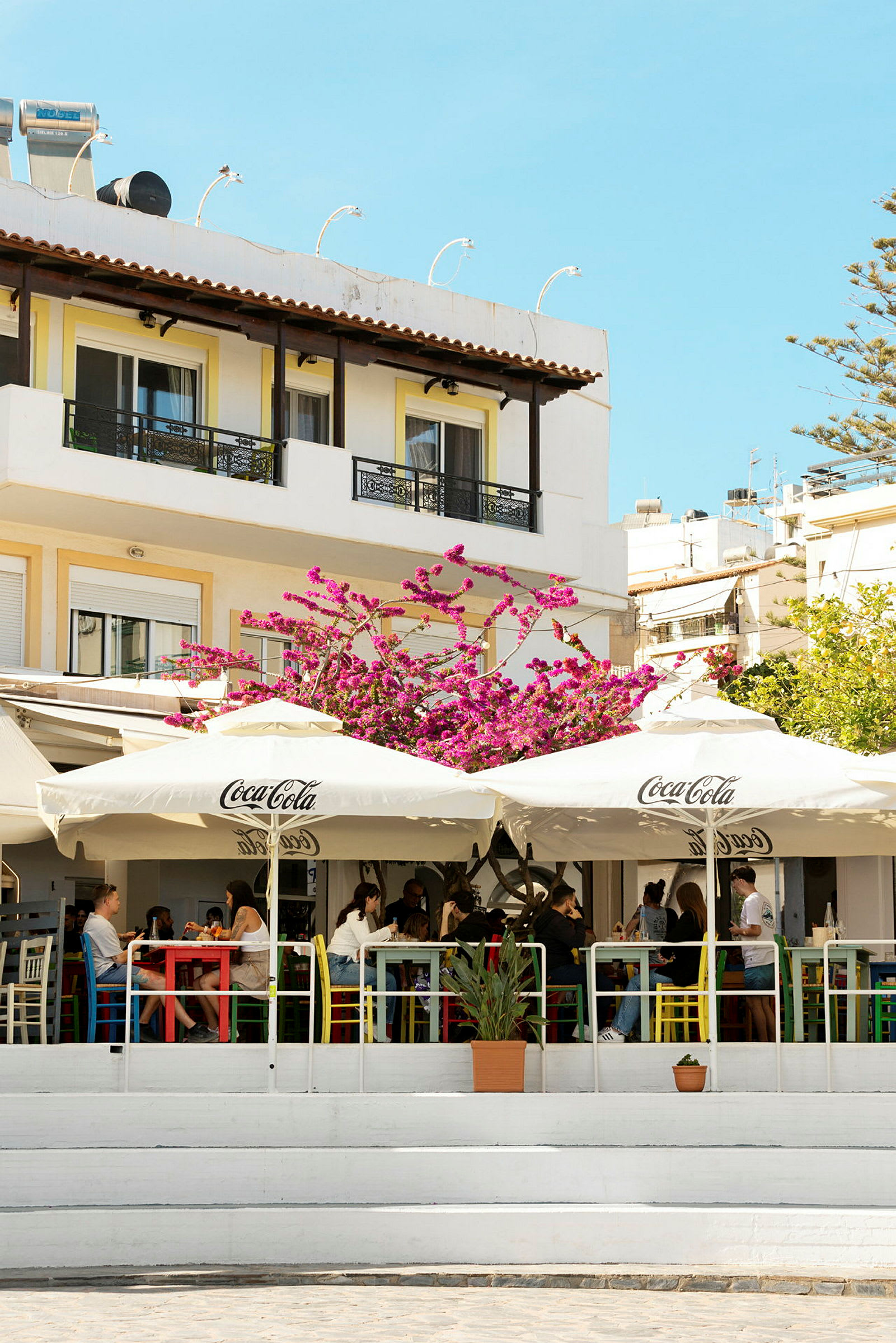 Udendørs café med parasoller og blomster foran en bygning. (AI-genereret tekst)