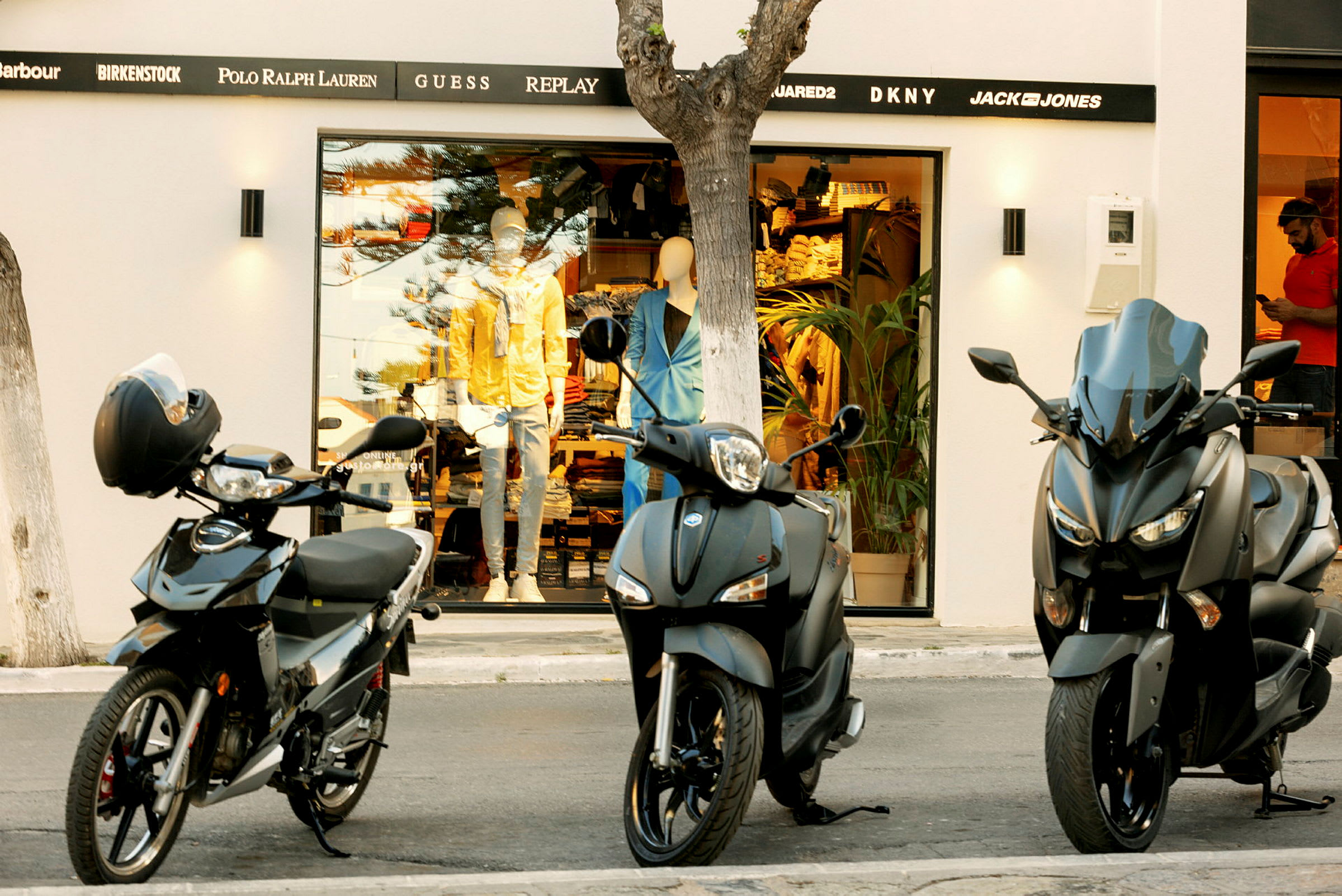 Tre motorcykler er parkeret foran en butik med et udstillingsvindue og mannekiner. (AI-genereret tekst)