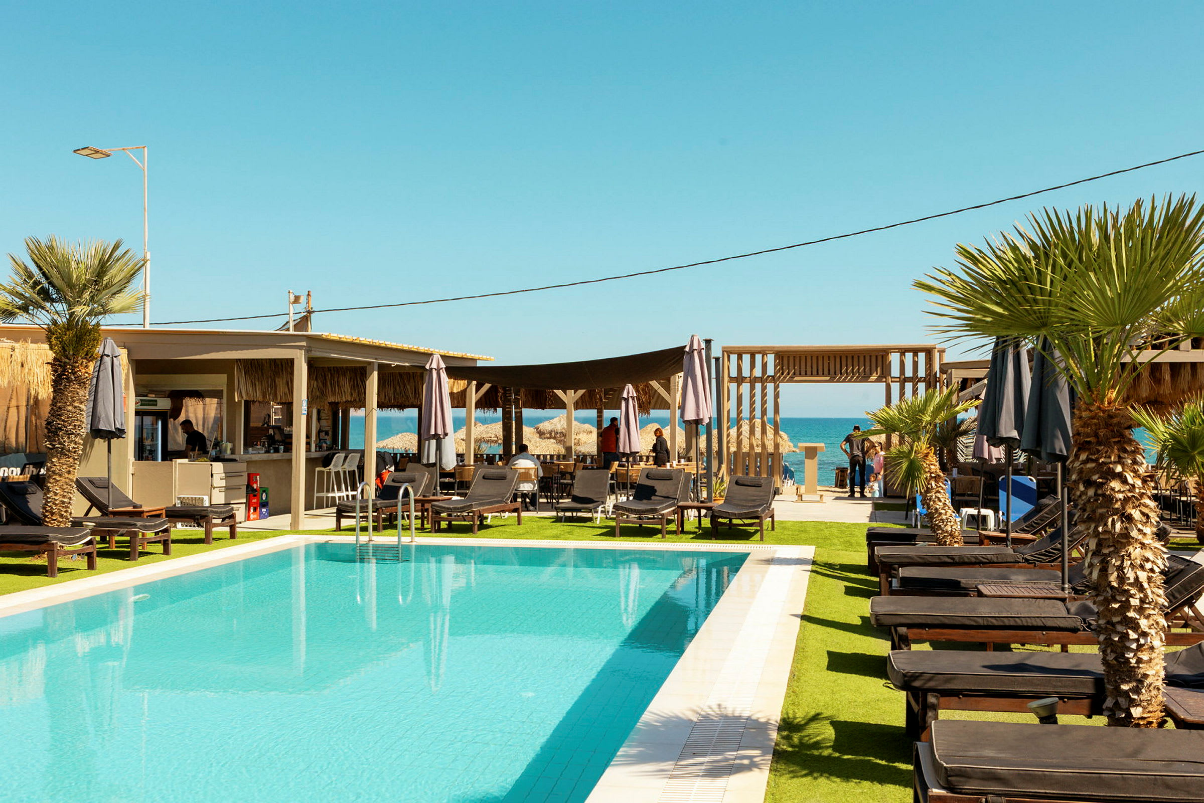 En swimmingpool omgivet af liggestole og parasoller med palmer og en serveringsbygning i baggrunden. I det fjerne ses havet under en klar blå himmel. (AI-genereret tekst)