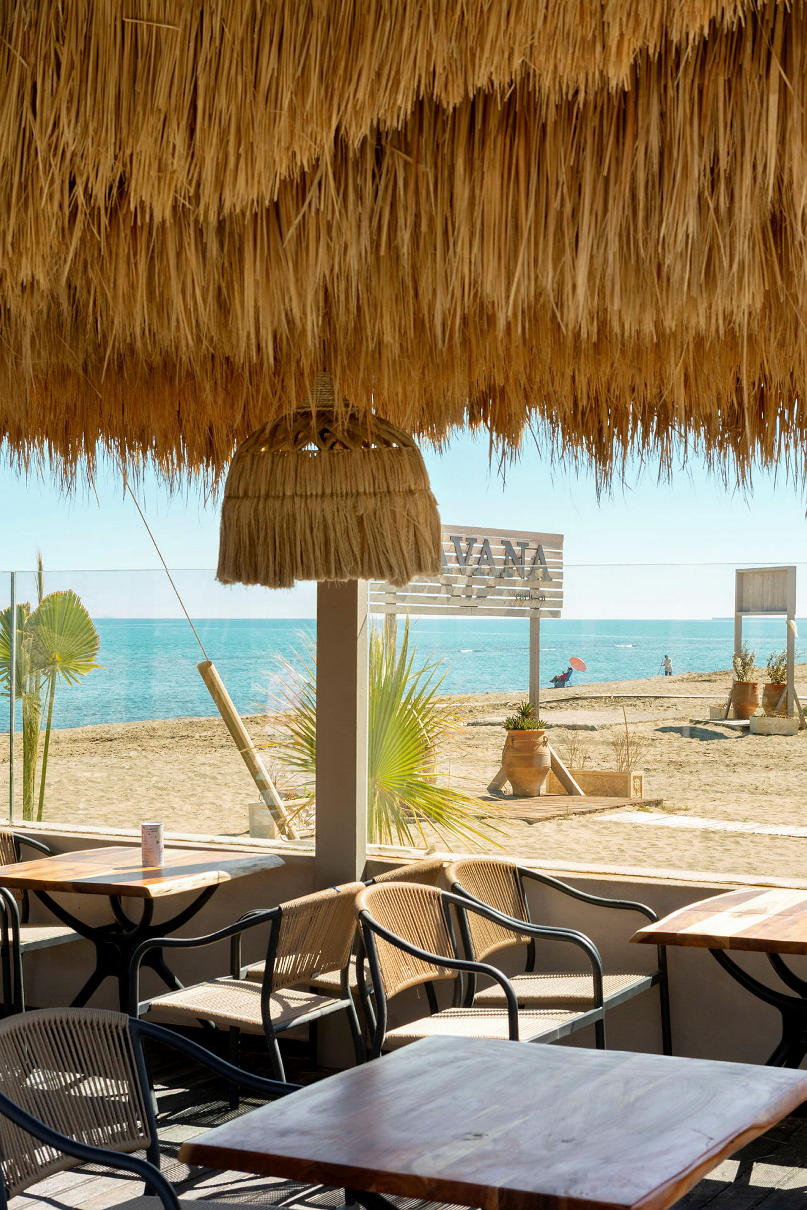 Udendørs serveringsområde med borde og stole under et halmtag med udsigt til en sandstrand og havet. (AI-genereret tekst)